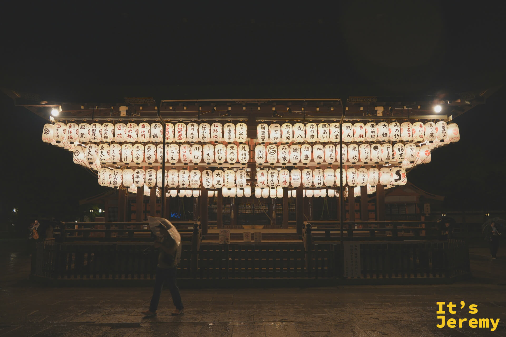 舞殿提燈全景，有撐傘遊客路過，雨夜氛圍最濃