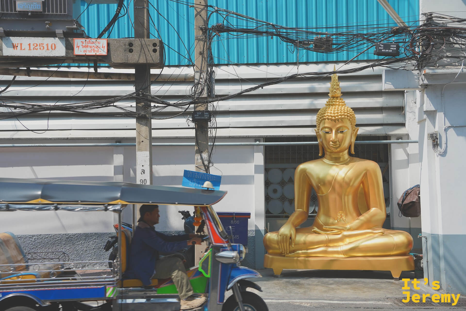 馬路邊一尊真人尺寸的金色坐佛被推到藍色 tuk-tuk 旁，準備被載走