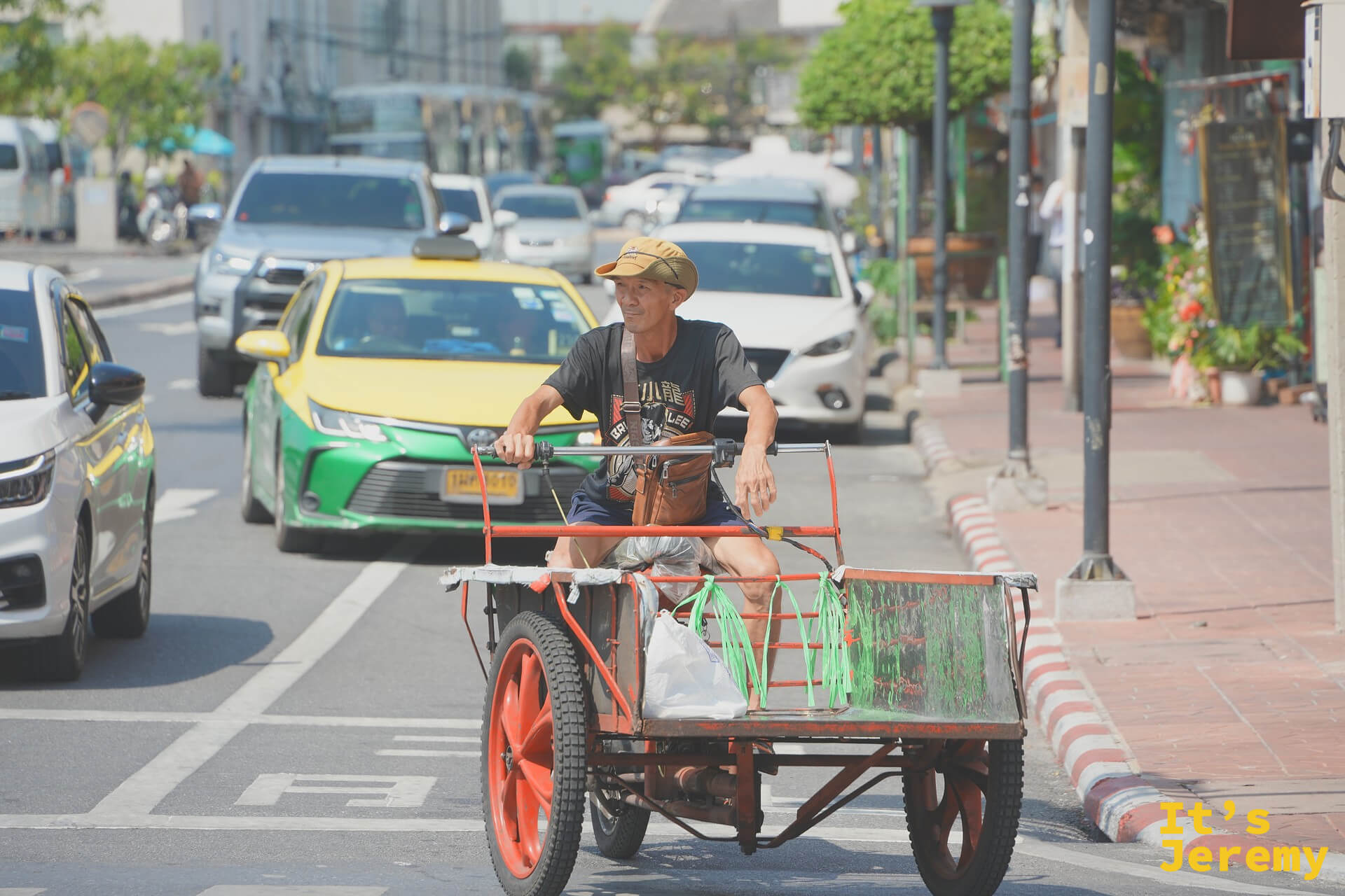 一位老先生戴著迷彩帽推著木板車過馬路，車上載著綠色塑膠袋——老城區最日常的搬運畫面