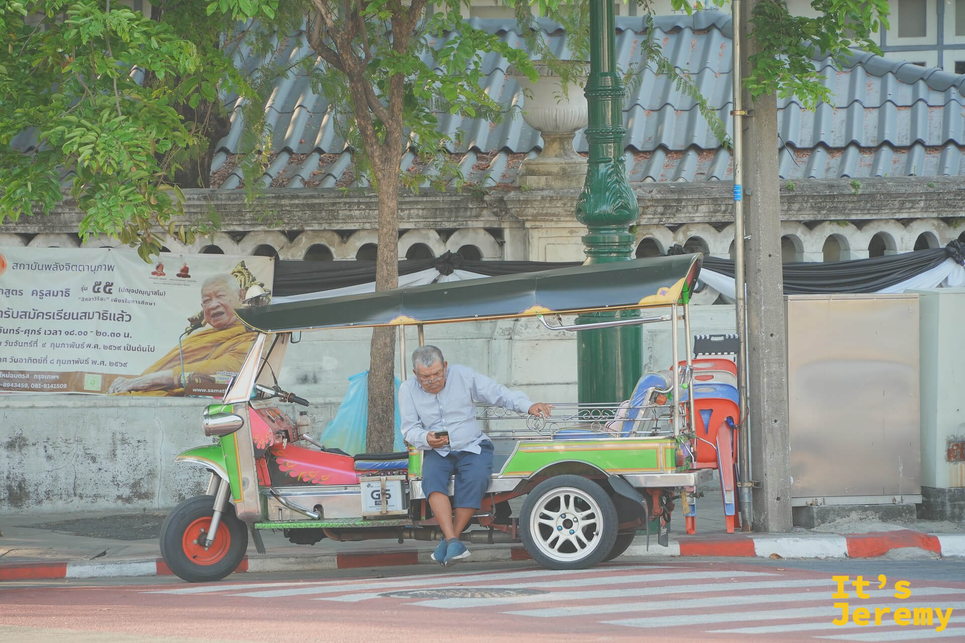 一輛停在運河邊的紅藍 tuk-tuk、司機坐在駕駛座上滑手機，後座掛著一張高僧海報