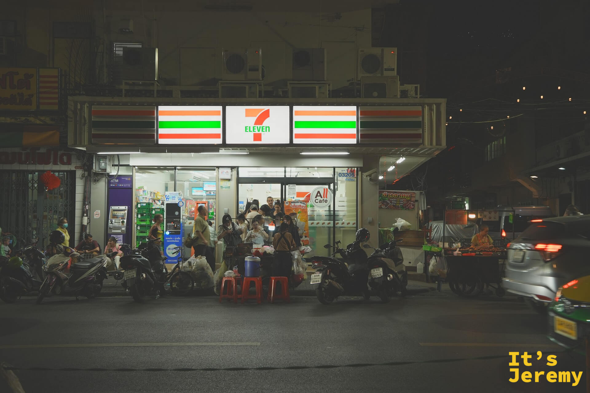 夜晚的 Tha Tien 7-Eleven，紅綠燈條把人行道切成兩半，門口擺著紅色塑膠凳的臨時小販