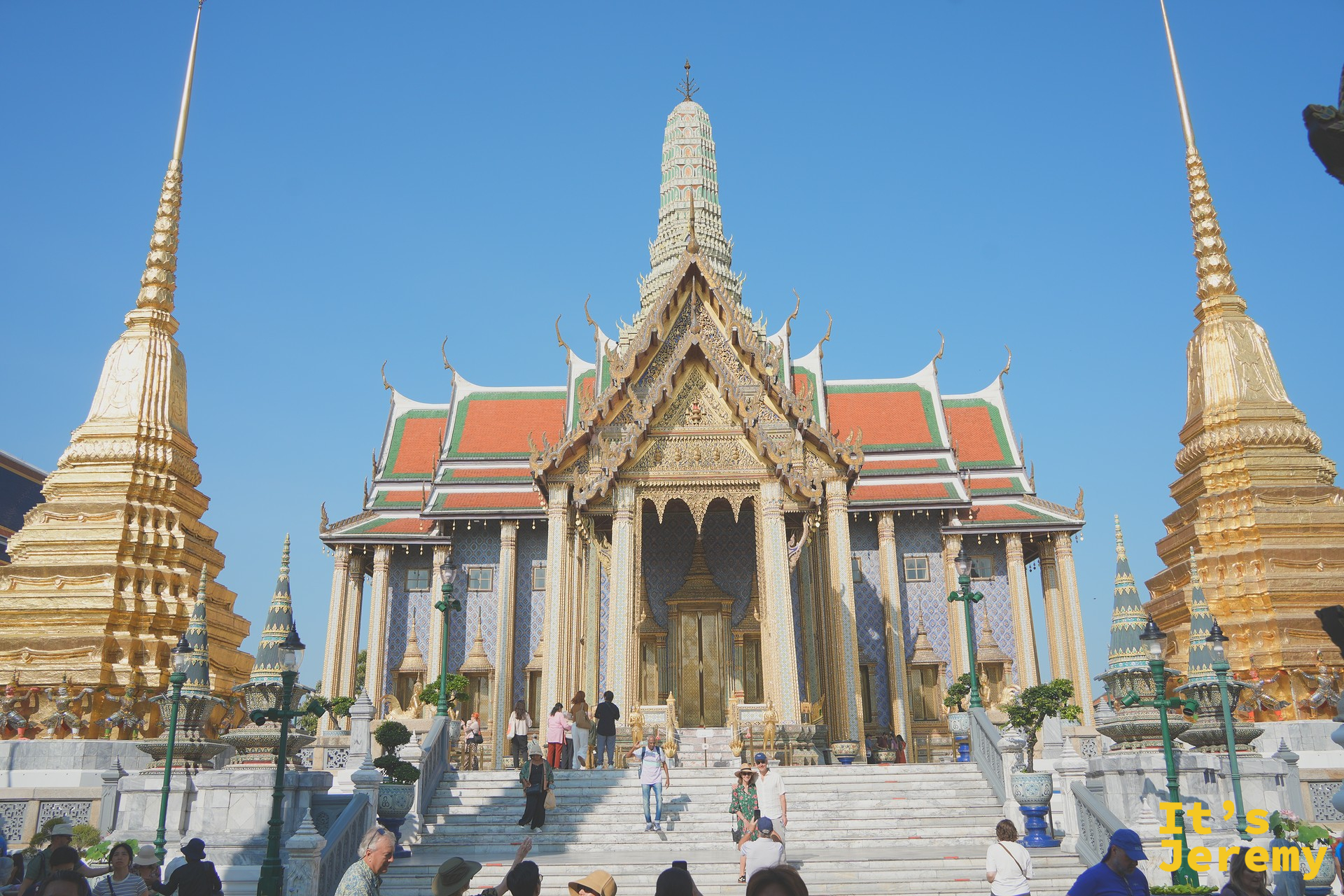 玉佛寺主殿正面全景，左右兩座金色 Chedi 與遊客上下台階
