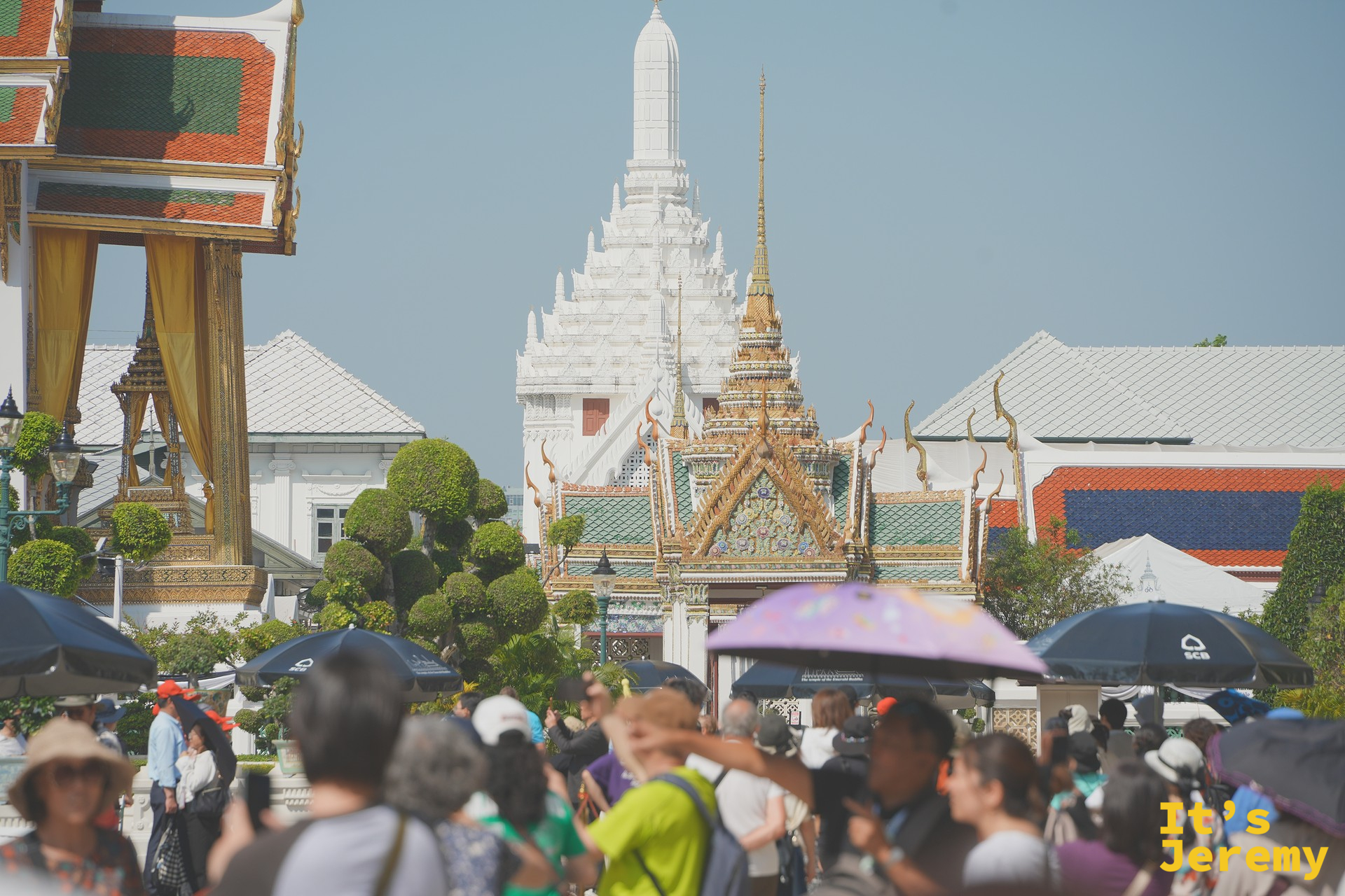 園區內擁擠的觀光人潮，遊客撐傘擋陽光，背景白色 Prasat 塔與金色屋頂