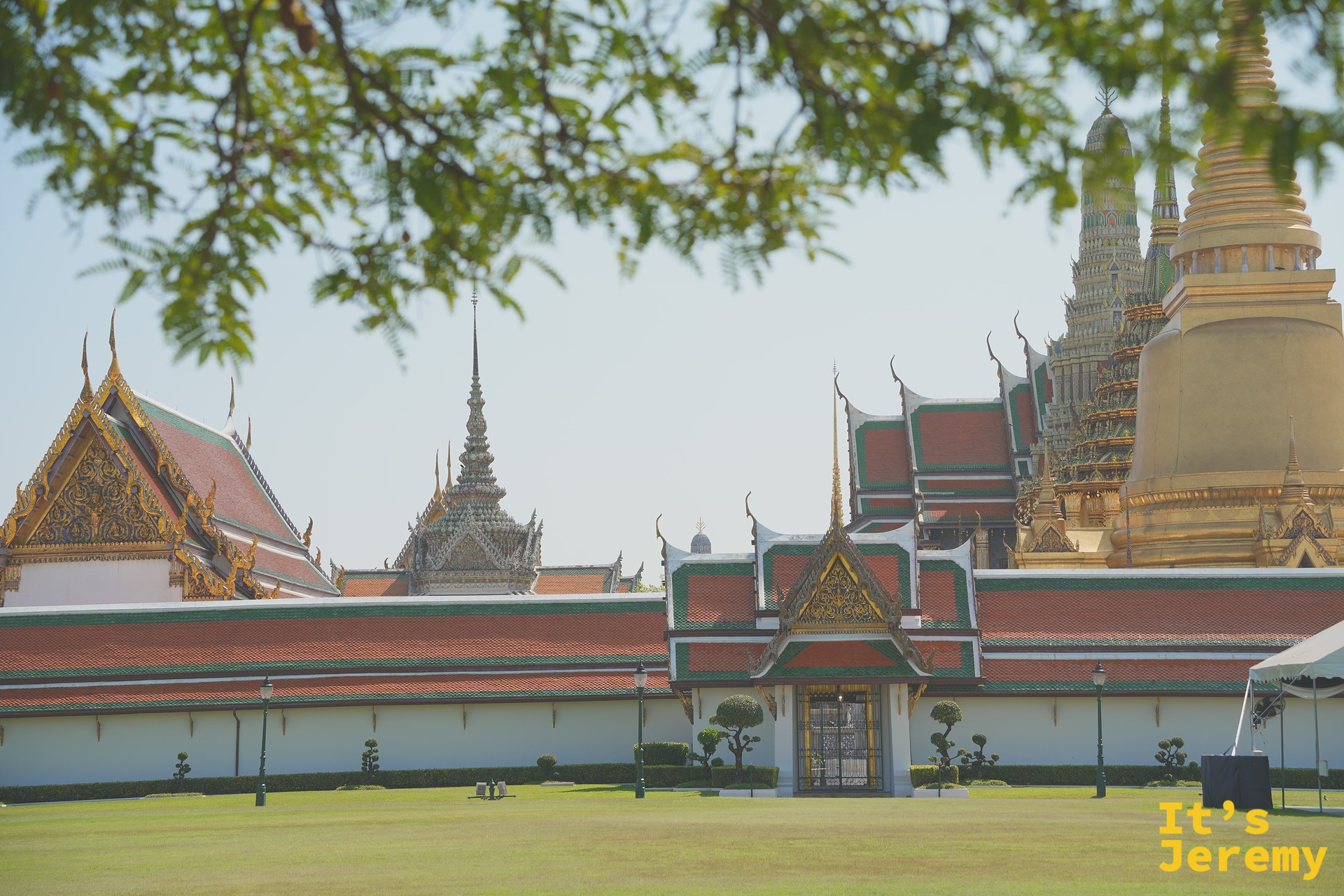 大皇宮外牆全景，樹蔭前景下的金色 Chedi 與紅綠瓦屋頂