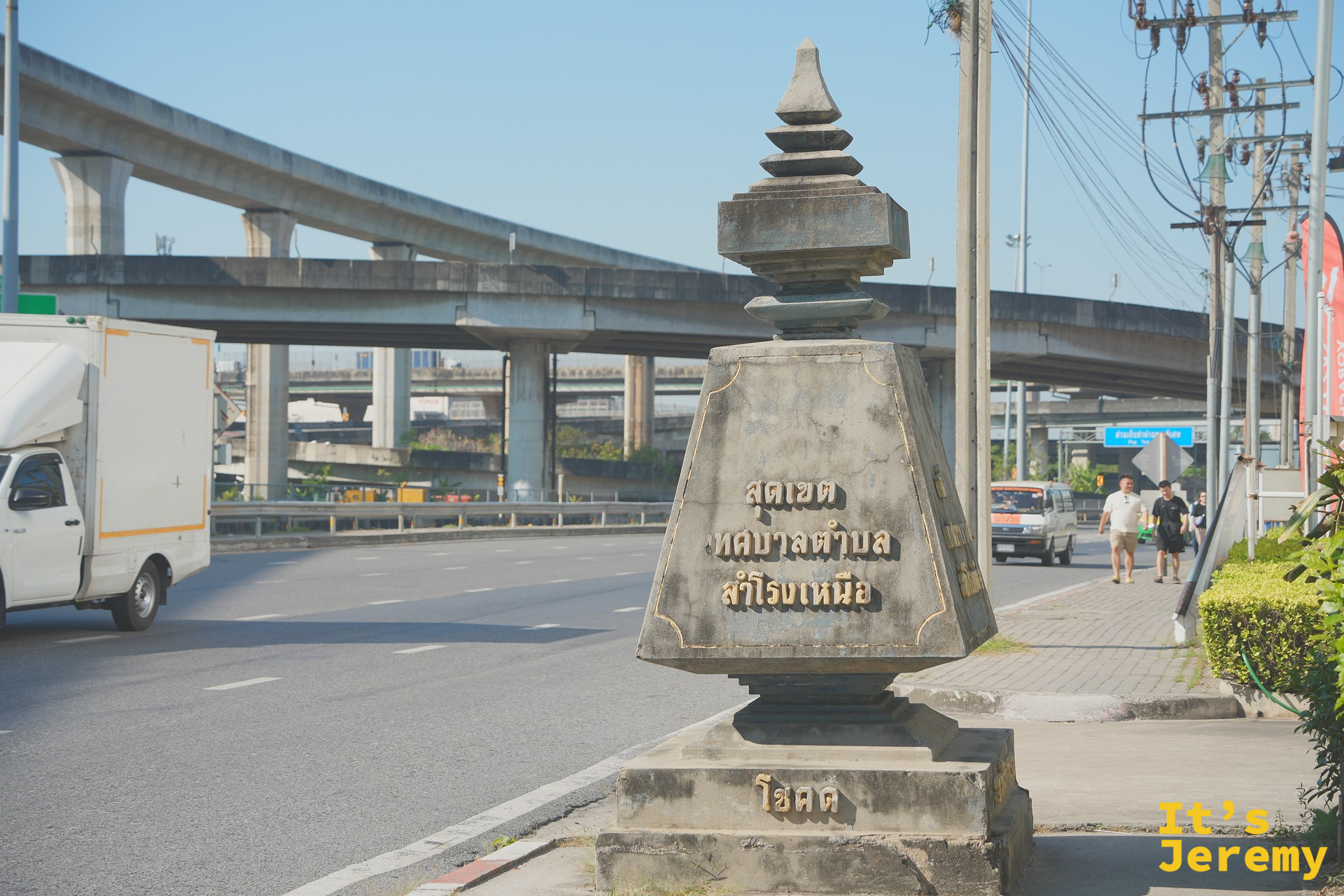 馬路邊的「Samrong Nuea 市鎮邊界」傳統石碑，背景是 BTS 高架軌道