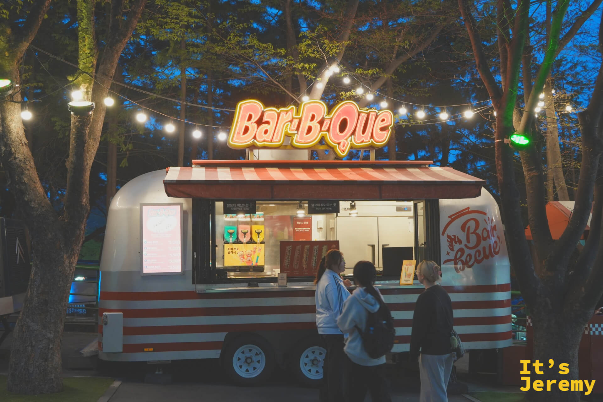 Bar-B-Que 餐車,銀白色復古車身、白底紅條紋帆布頂篷,車頂掛著黃紅色霓虹「Bar-B-Que」招牌,三位女生站在窗口前點餐,車頂兩側拉著一串小串燈