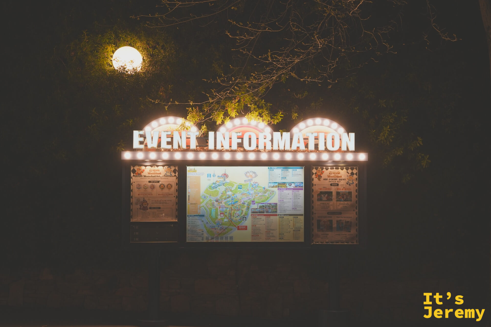 Event Information 公告牌:頂部三個拱形邊框鑲滿黃色小燈泡,下方三塊看板印著園區地圖與當日活動表,背景是深色樹影與一盞發光的球形路燈