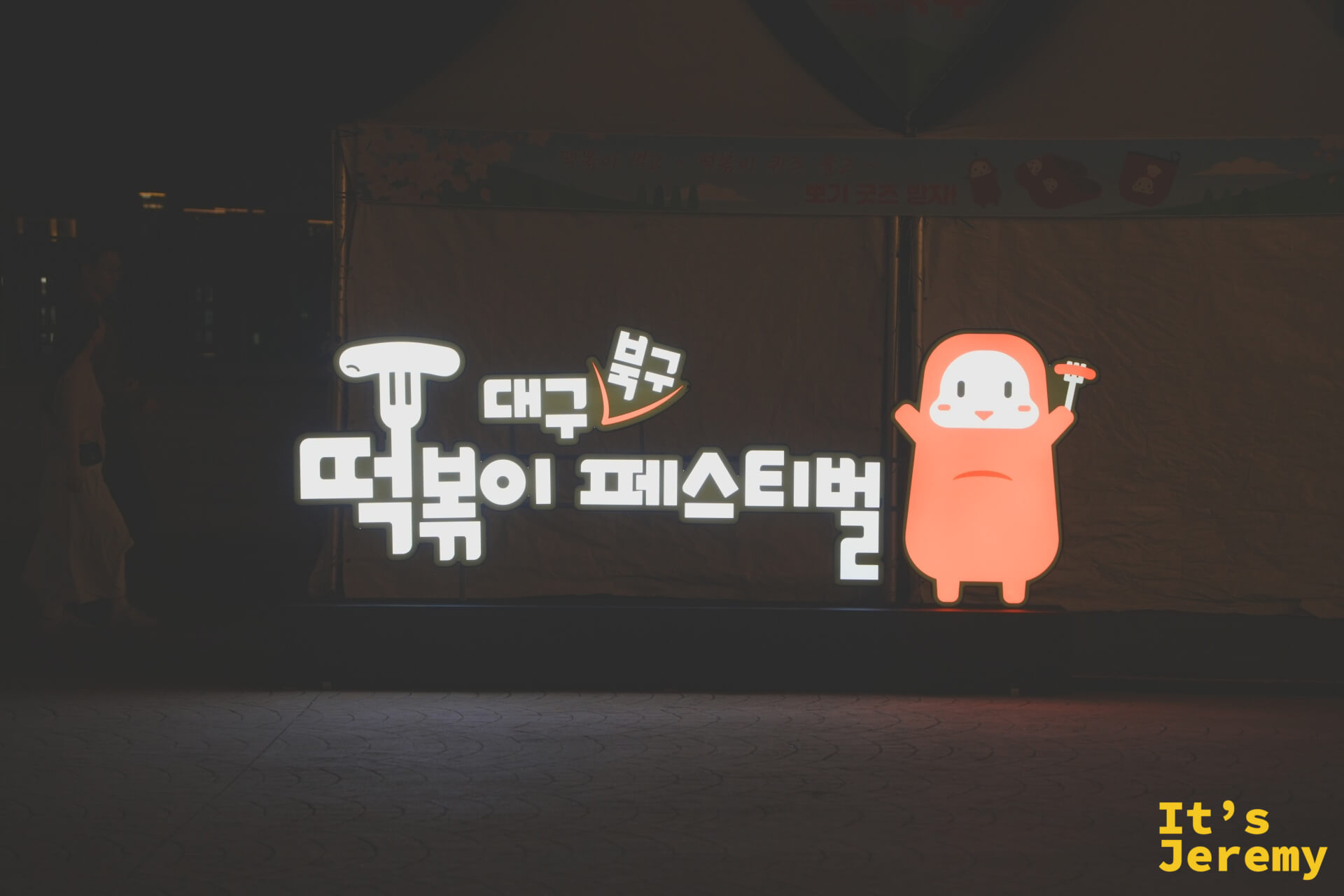 大邱북구떡볶이페스티벌(大邱北區辣炒年糕節)的發光招牌,黑色帳篷前的白底字樣與右側橘色卡通吉祥物(手拿叉子與年糕)發光跳出畫面