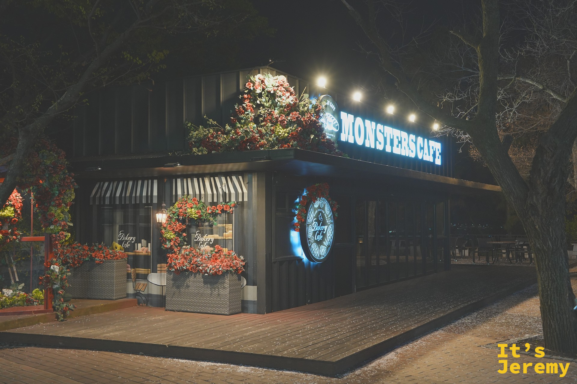 Monsters Cafe 外觀,黑色貨櫃屋造型咖啡店,藍色霓虹「MONSTERS CAFE」招牌,門口被大量紅花綠葉裝飾包圍,圓形「Bakery Cafe」標誌掛在門邊