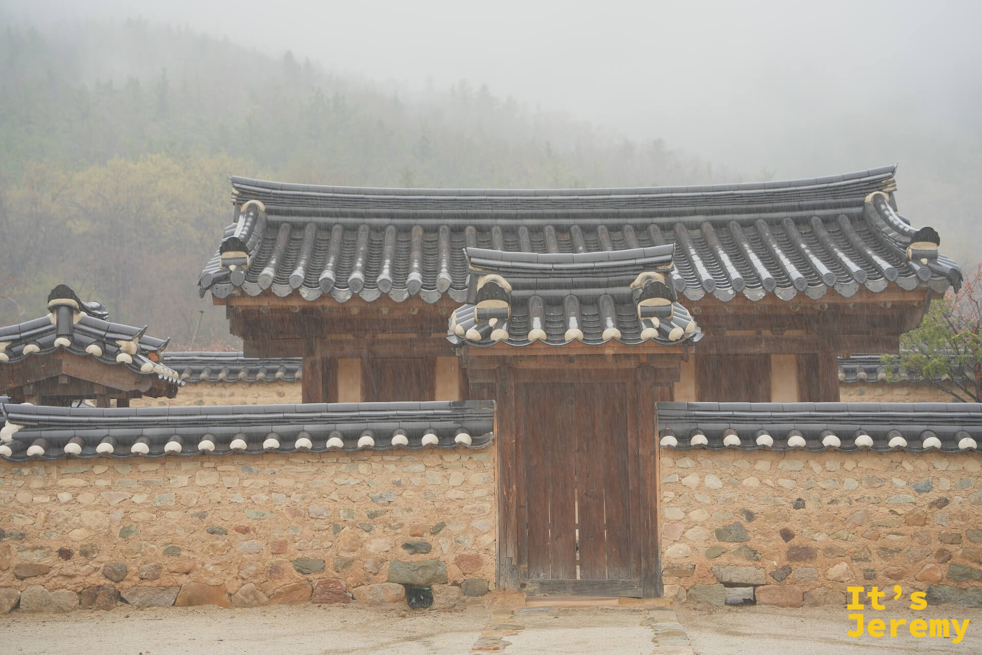 雨中的漆谷韓屋村正門，青瓦大門後是霧氣瀰漫的山，雨絲清晰可見