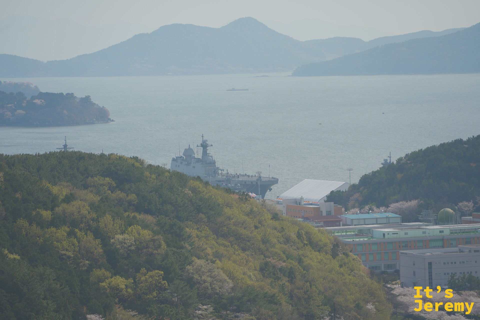 港灣後方山丘間停泊的韓國海軍艦艇,與綠色山坡和春日粉白花樹形成強烈對比