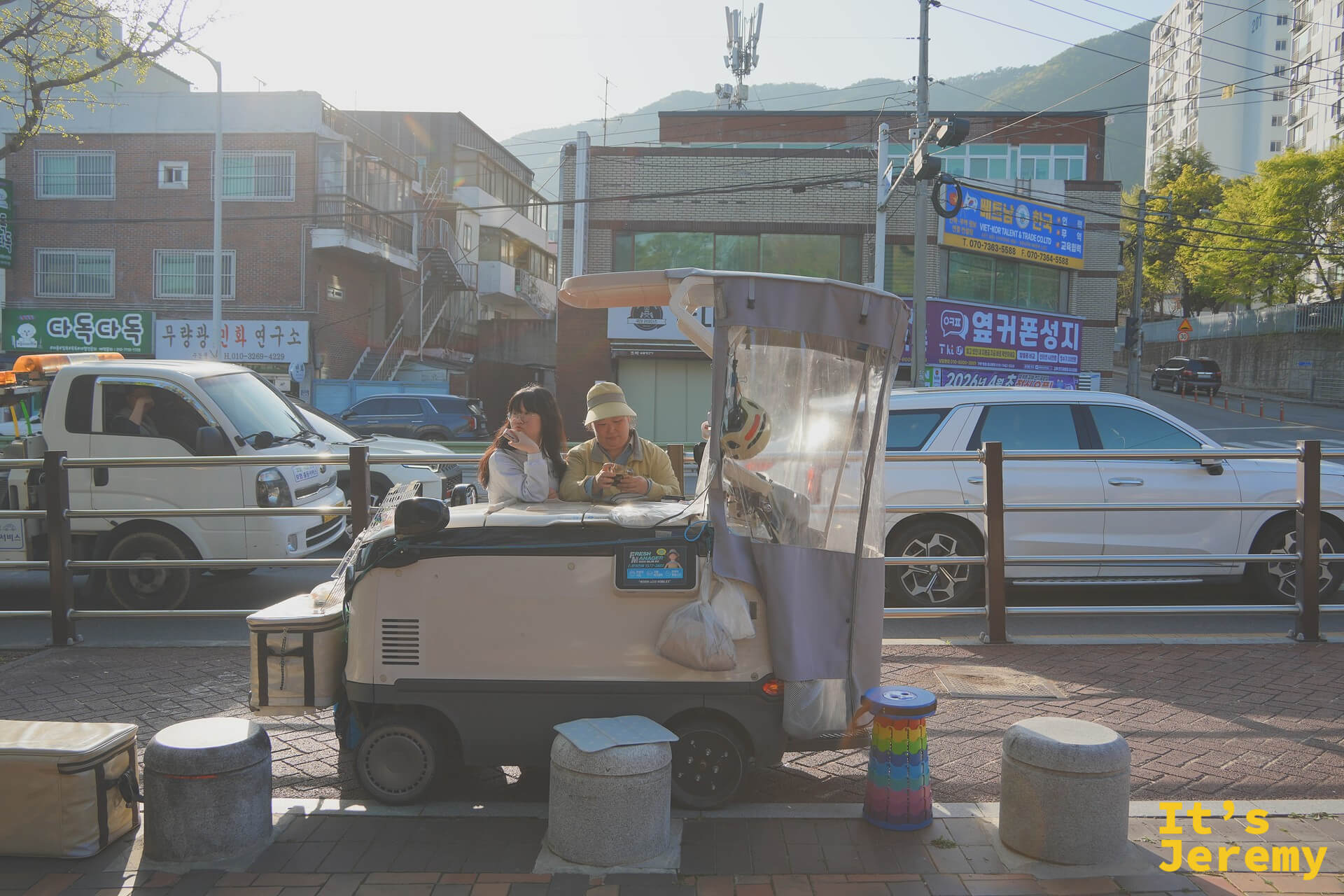 公園外圍街道旁，一輛改裝餐車停在路邊，攤主和女孩並肩在車內，春日斜陽把整個畫面染成金黃