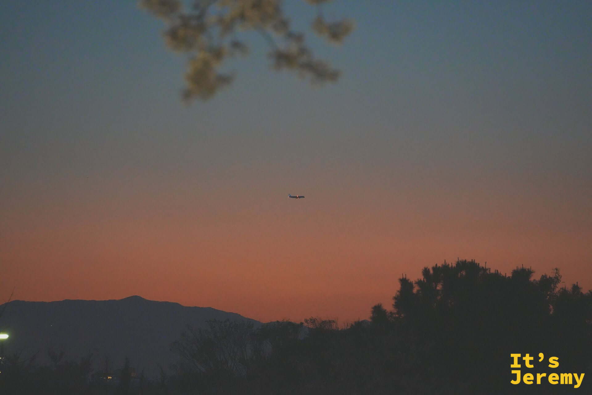 夕陽餘霞染紅整片天空，遠處的山稜線壓低成剪影，一架飛機悄悄穿越橘色的天幕