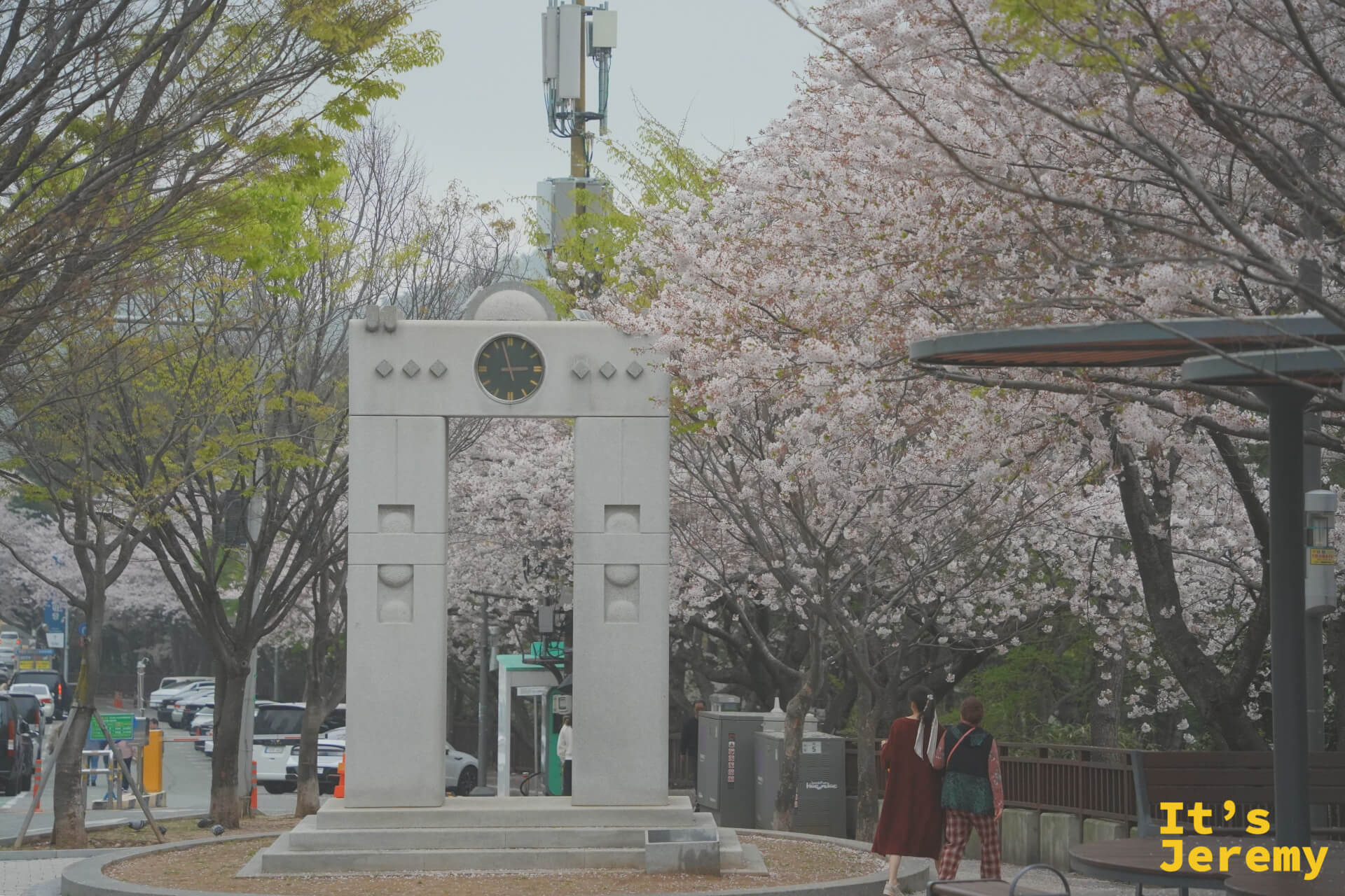 石製時鐘拱門矗立於迎月路公園入口，兩側吉野櫻盛開，一名穿著韓服的旅客從拱門前走過