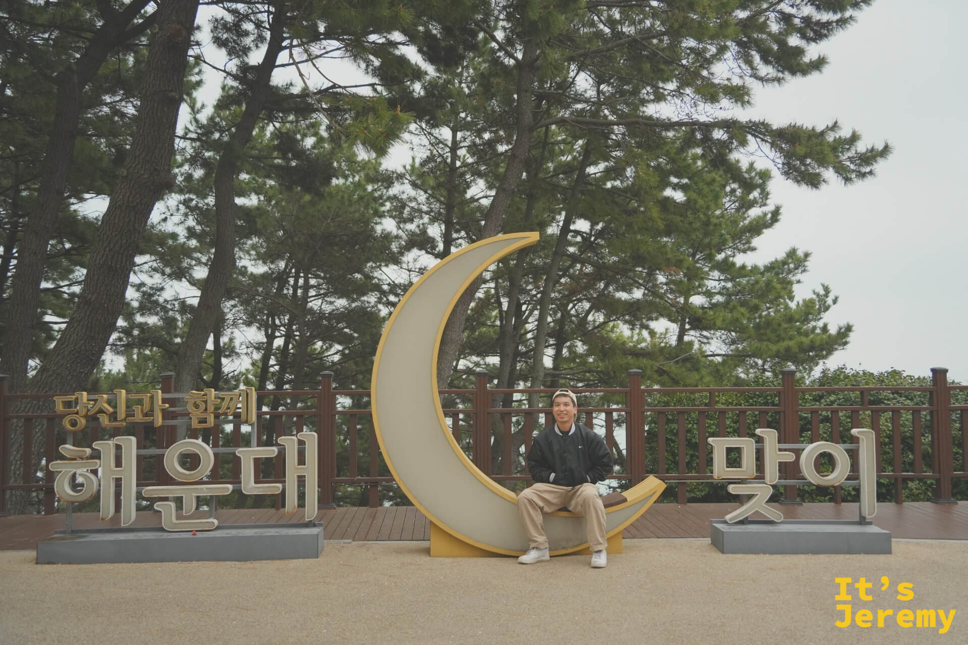 Jeremy 坐在黃色月牙形地標的凹槽上，背後是茂密的松林，天色微陰