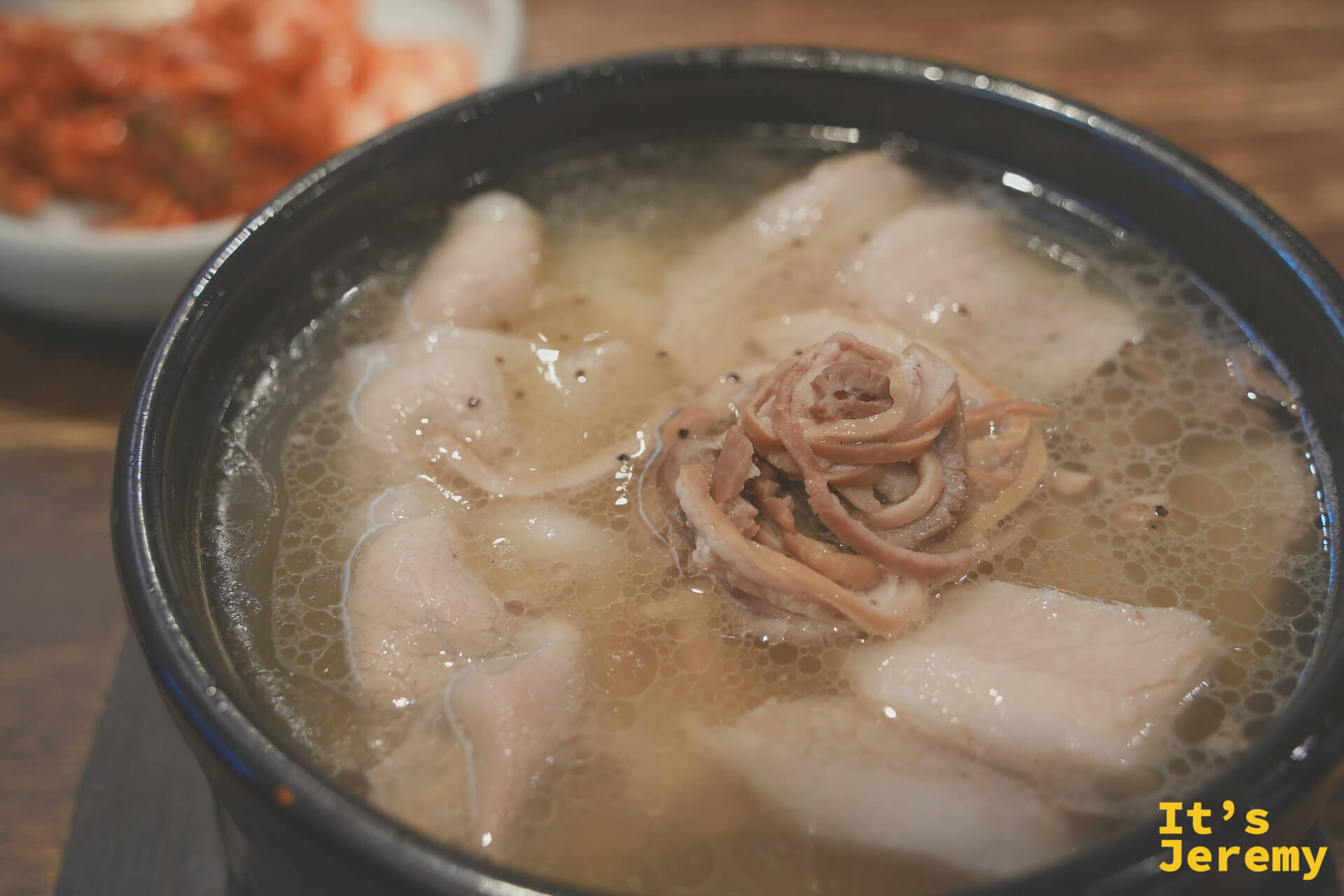 釜山式豬肉湯飯近景，清亮湯底中漂浮著厚切豬肉片與捲曲的豬橫膈膜，後方是泡菜小碟
