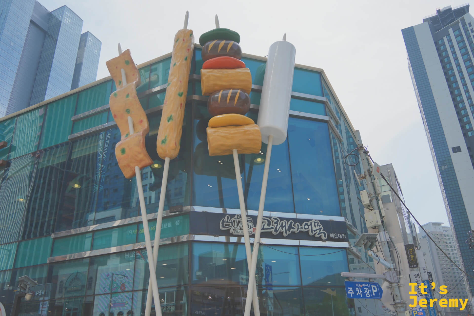 從路口望向古來思魚板海雲台店，三根巨型魚板串矗立街角，海雲台鬧區高樓為背景