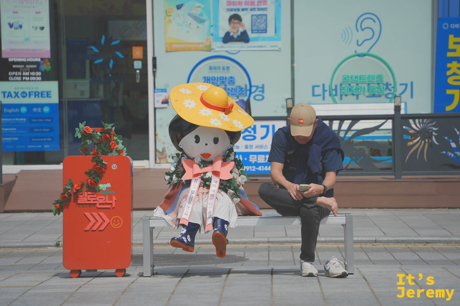 海雲台街頭,穿著傳統韓服、戴著黃色大草帽的可愛偶人裝置坐在長椅上,旁邊的男子低頭滑手機;這種人與裝置共存的街頭畫面,是釜山日常裡意外好拍的素材