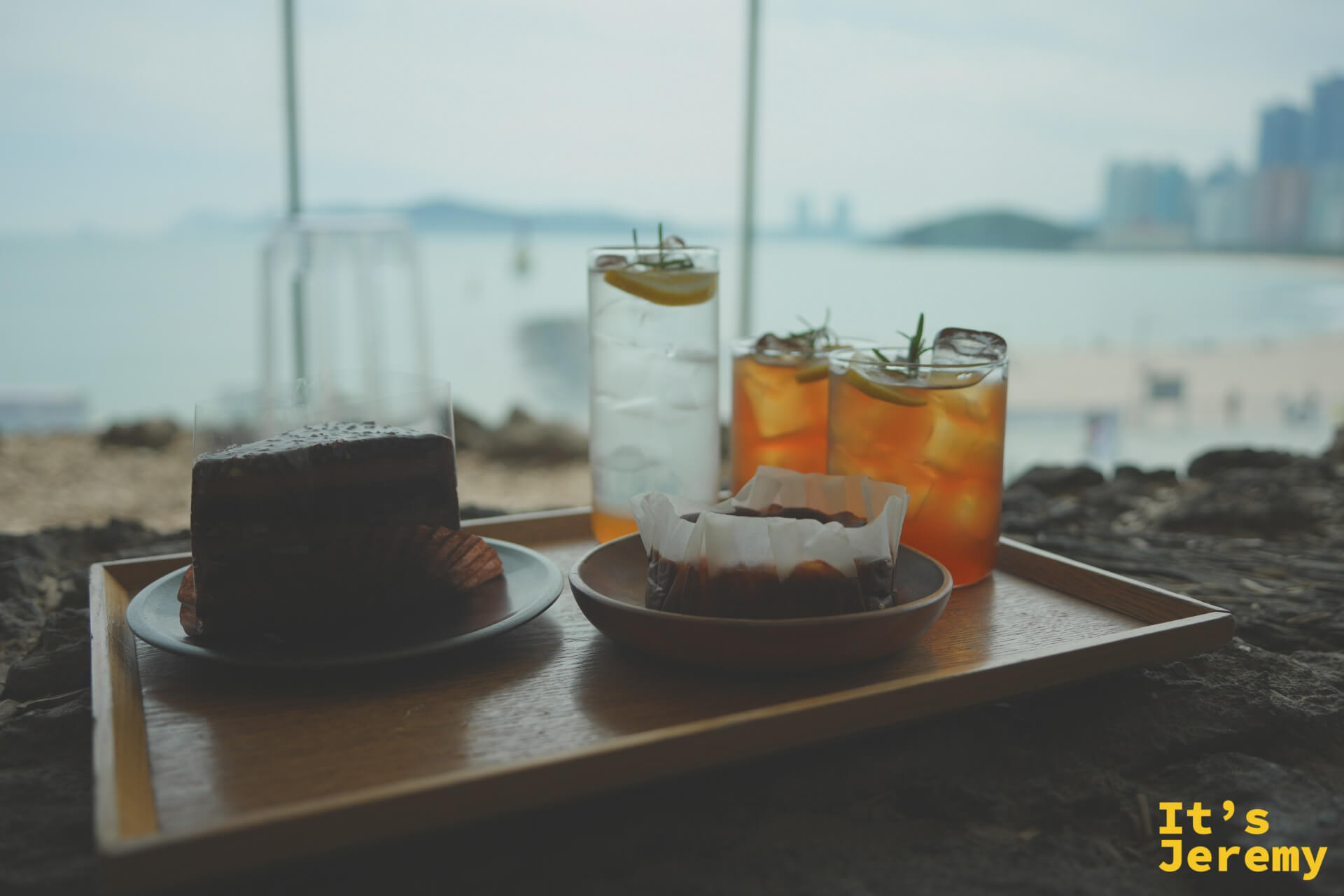 木托盤上的巧克力蛋糕、巴斯克乳酪蛋糕與三杯冰飲，背後是模糊的海岸線