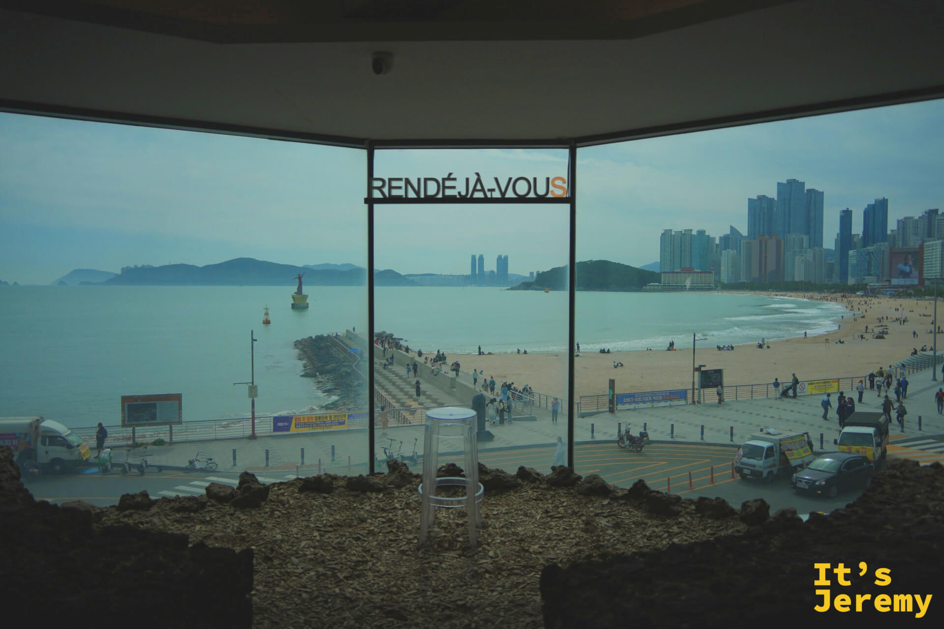 RENDÉJÀ-VOUS logo 懸掛在落地窗鐵框上，窗外是海雲台防波堤全景