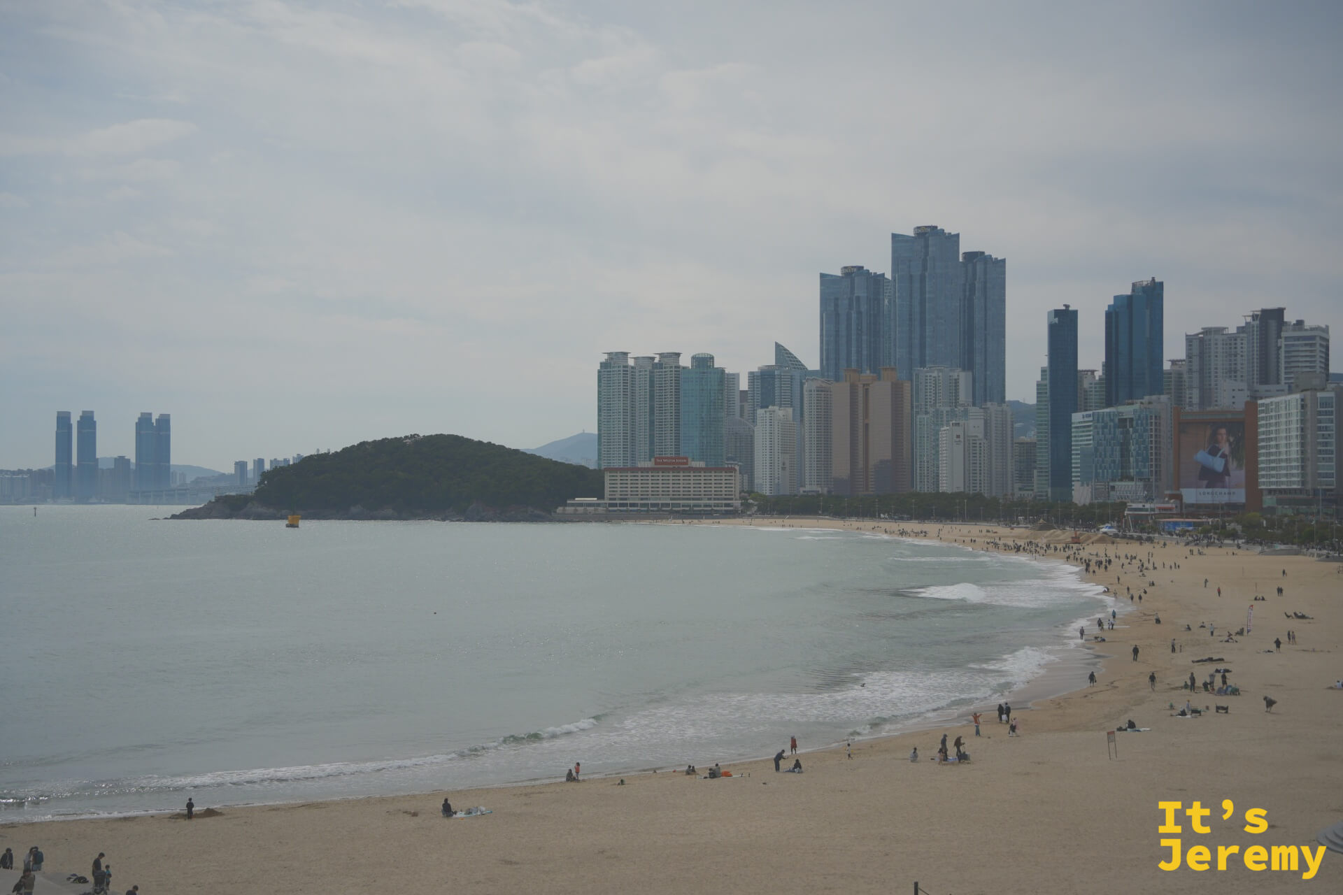 海雲台全沙灘弧線，從防波堤望向冬柏島方向，高樓與海灘交疊