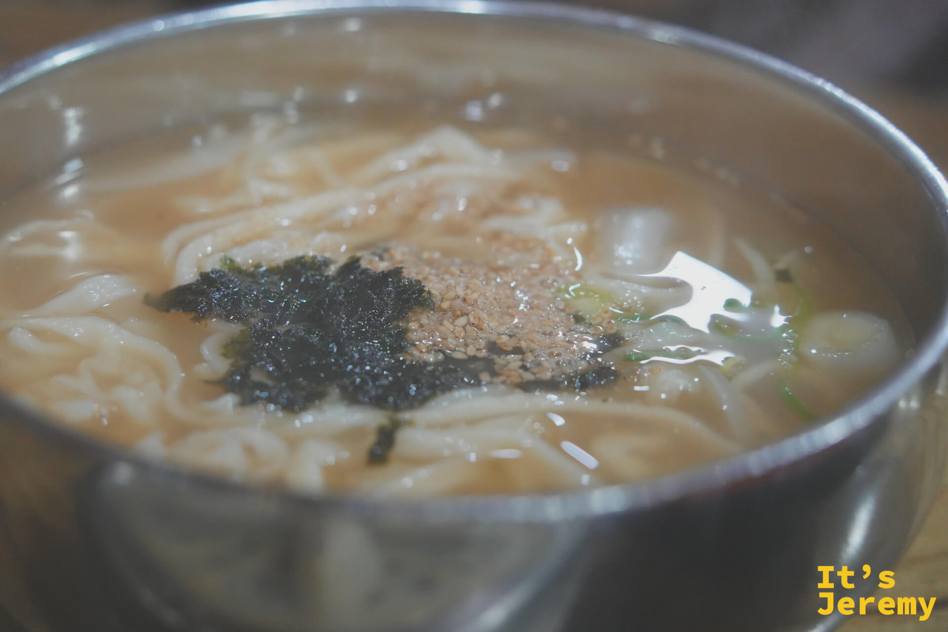 칼국수的近景:清澈骨湯、寬厚手切麵條,頂上撒著海苔碎與白芝麻