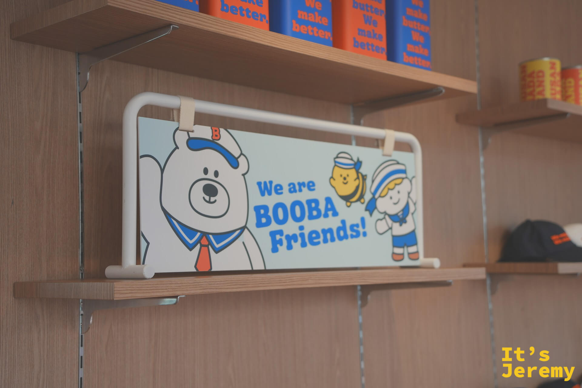 店內木架上的「We are BOOBA Friends!」角色插畫板，BOOBA 熊、蜜蜂和水手女孩三人齊聚，架上排著藍橘配色商品罐