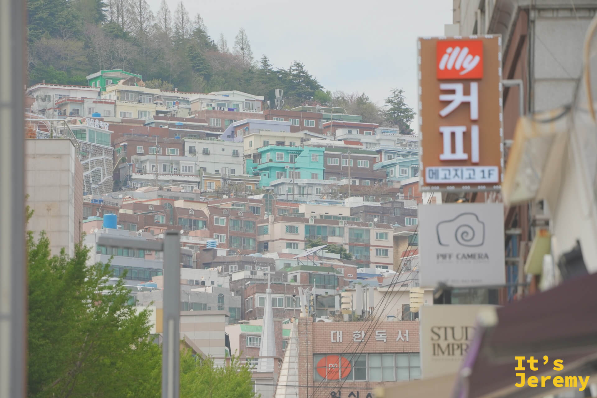 從街道往上望，山坡上密密疊疊的釜山住宅，前景是 illy 咖啡招牌和「커피」大字