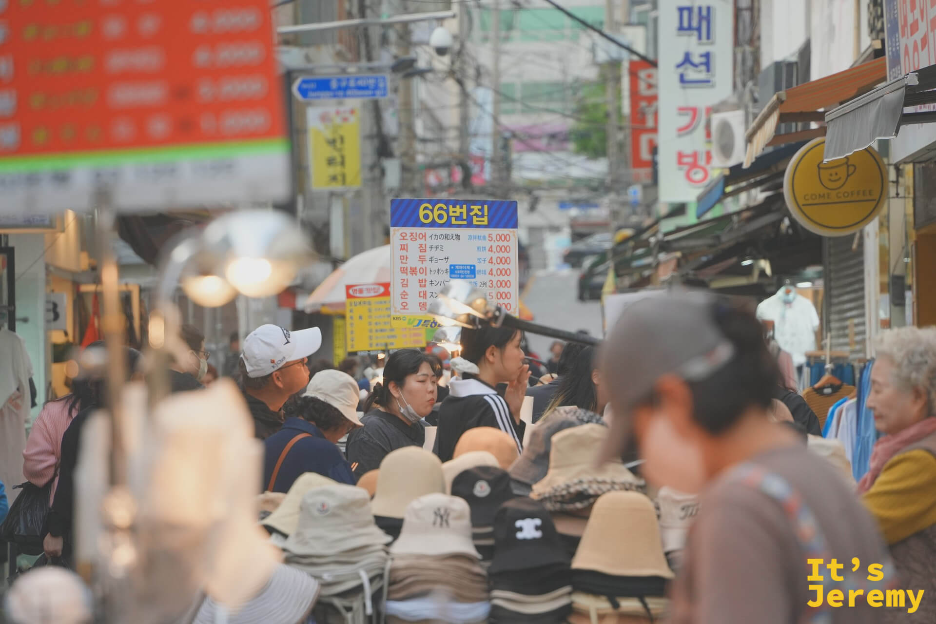 國際市場巷弄人潮，帽子攤、街食、招牌密密疊在一起，66번집的菜單牌懸在人群上方