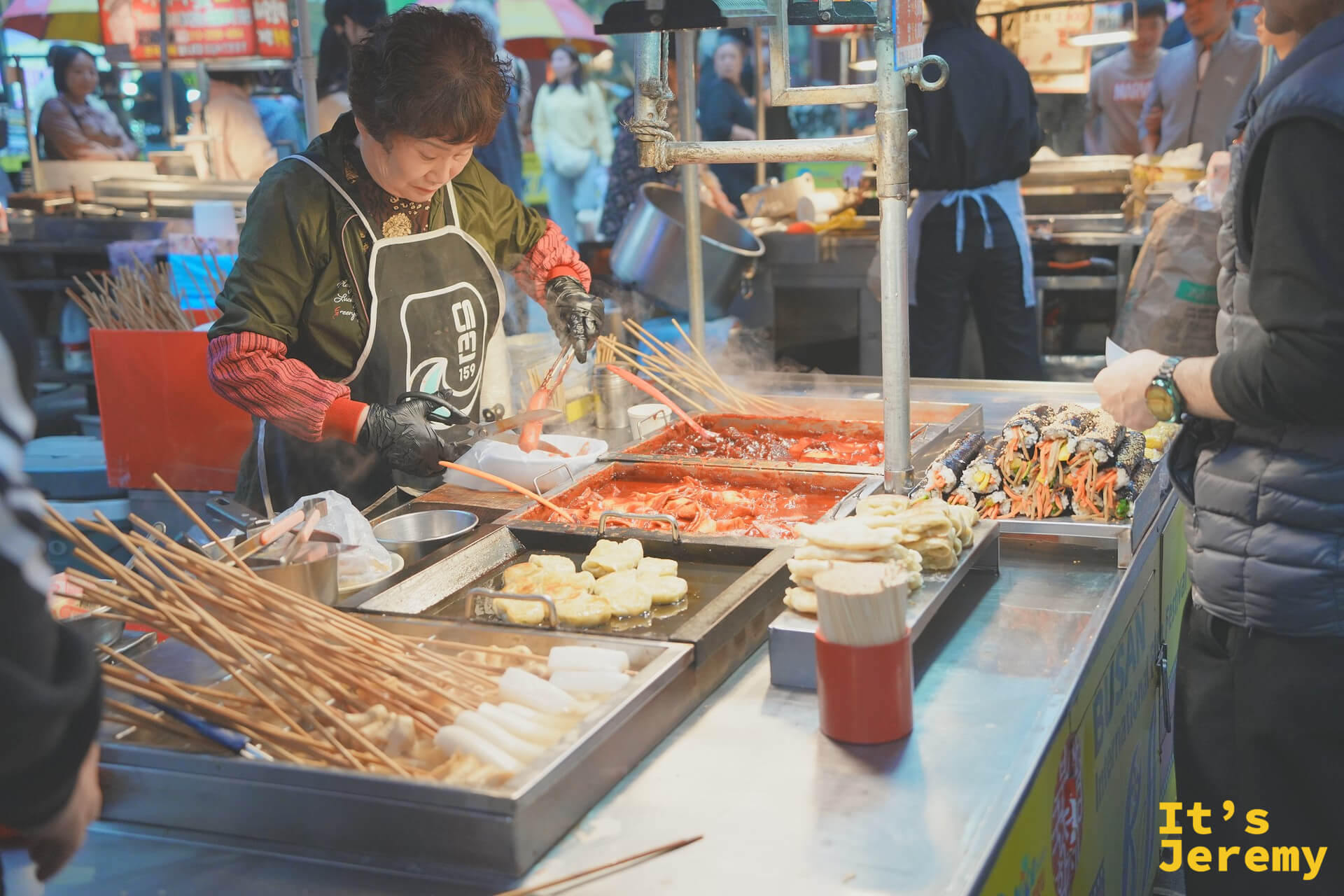 國際市場街食攤女老闆翻炒鐵板上的年糕、煎餃與海帶飯卷，熱氣四散