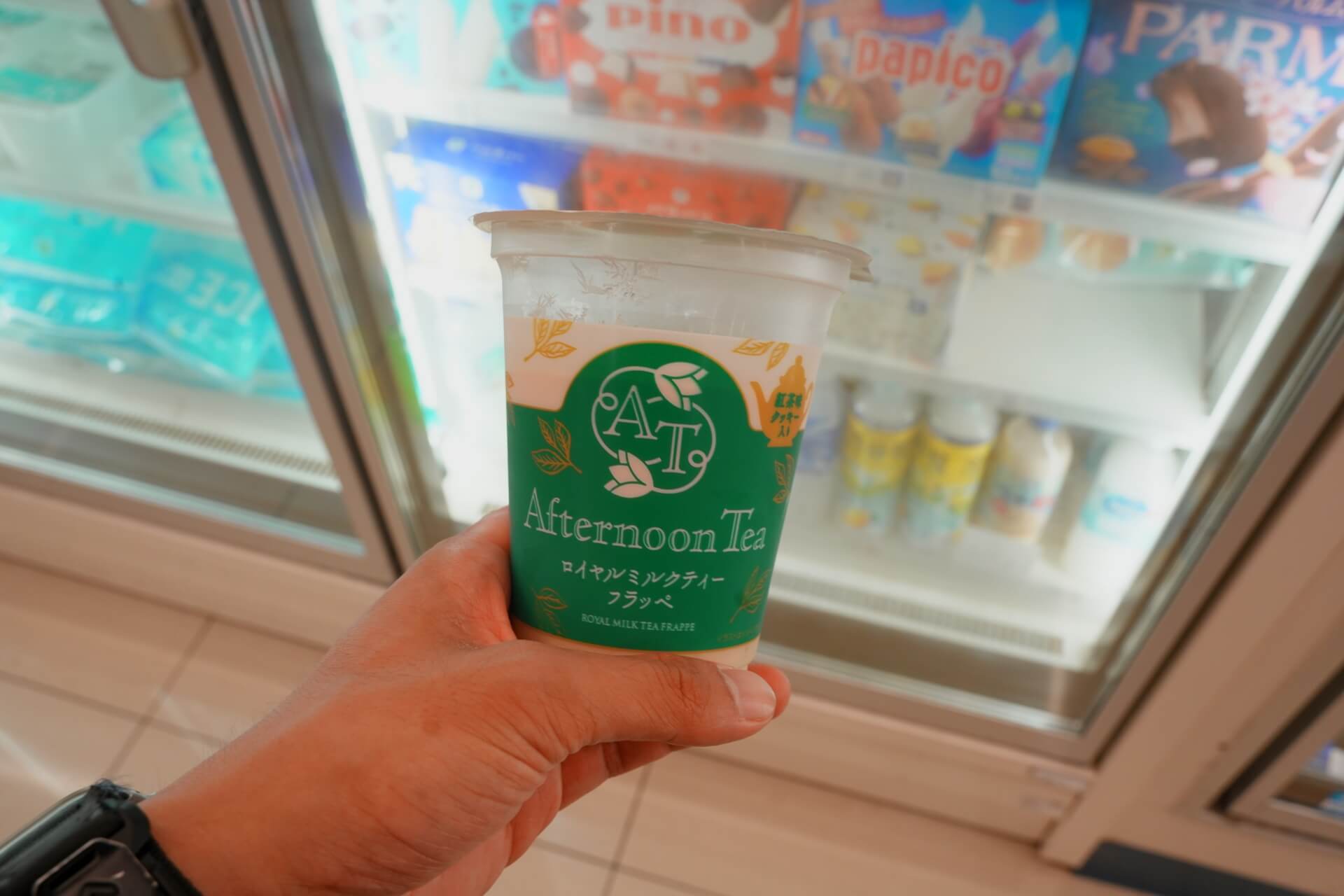 在全家冰箱前手持 Afternoon Tea 聯名「ロイヤルミルクティーフラッペ」，背景是各式冰品