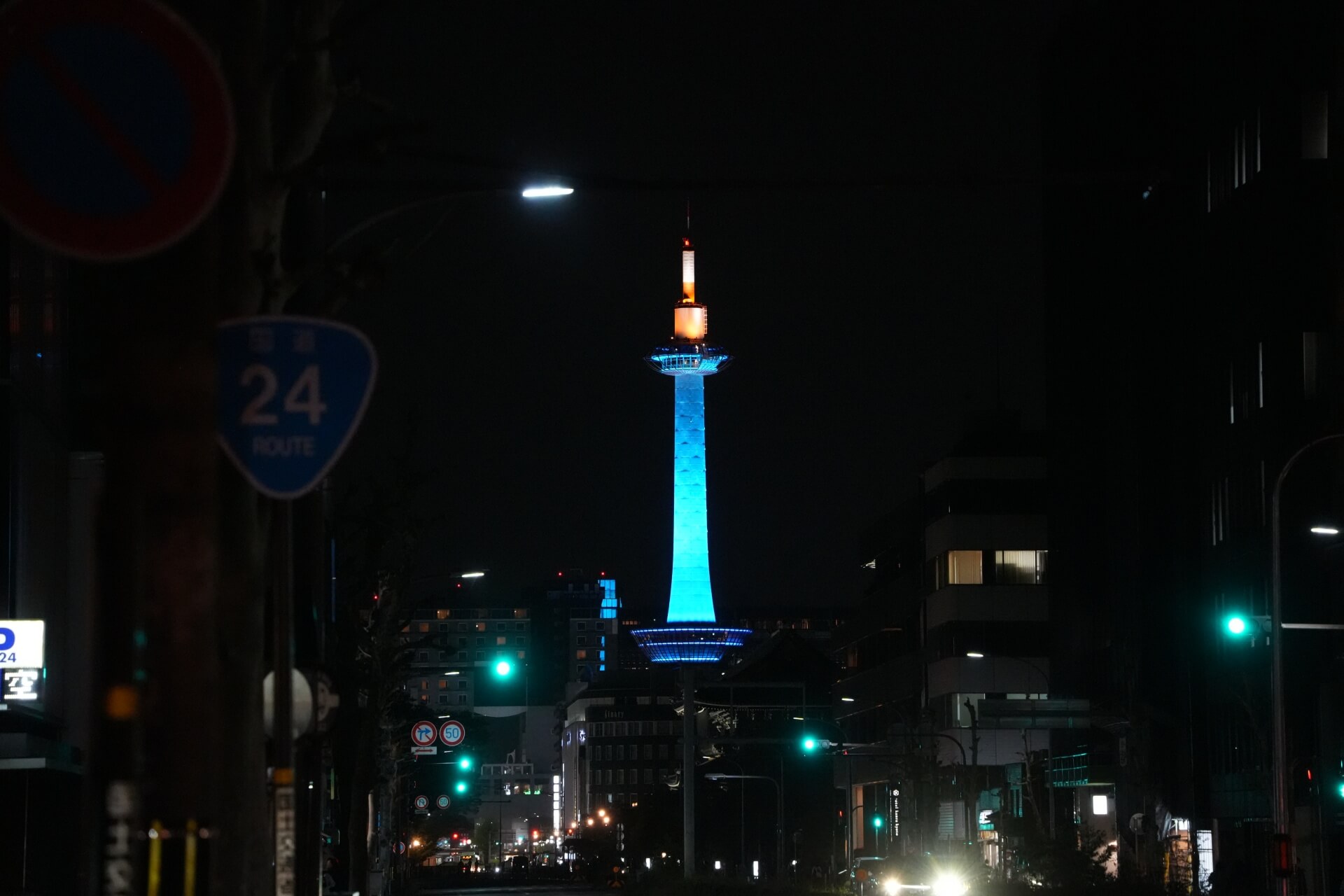 入夜的京都街道，京都タワー以藍色燈光點亮，從街道盡頭的空曠處正面入鏡