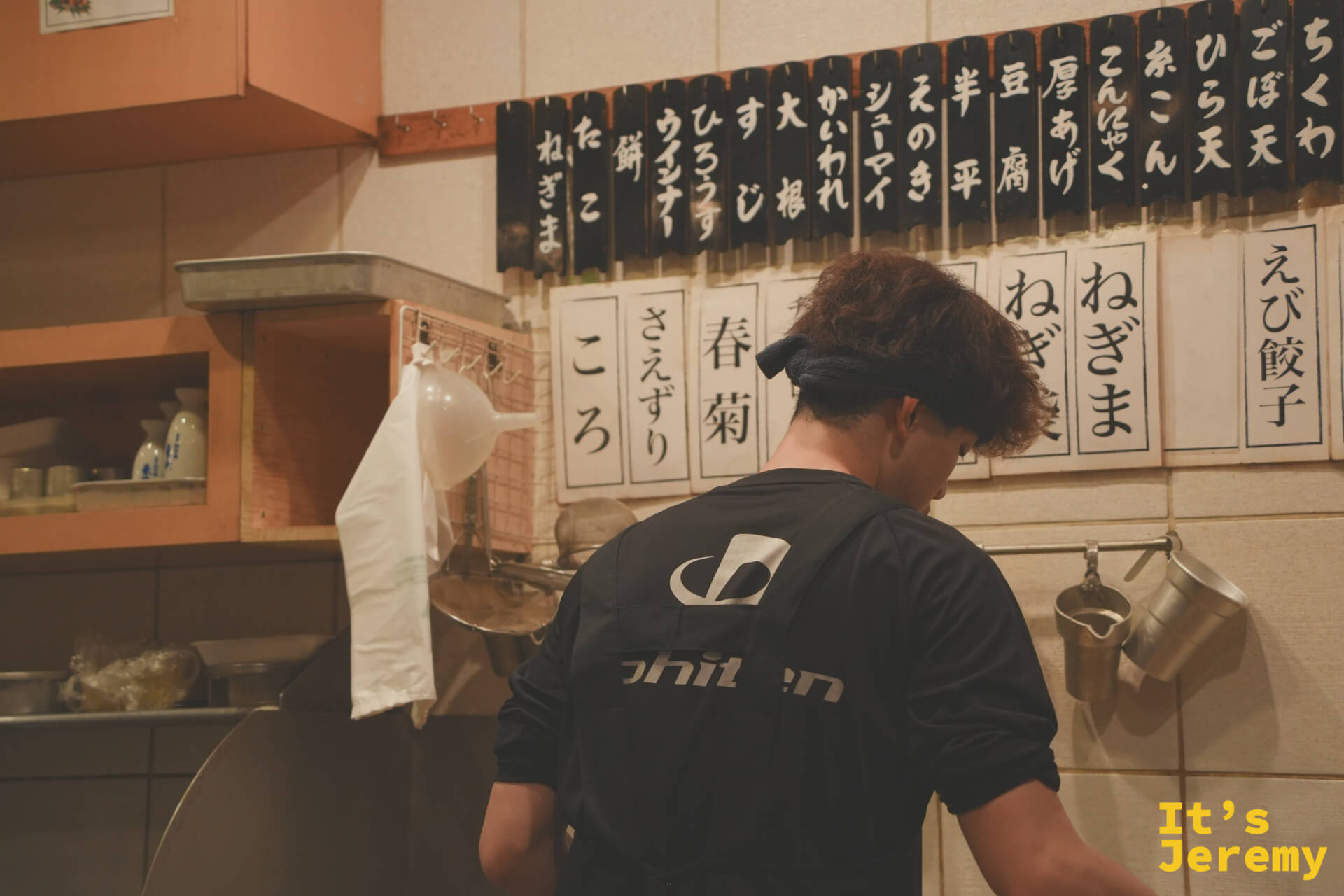 師傅背對鏡頭作業，身後牆上貼著印刷字卡，列出所有おでん品項