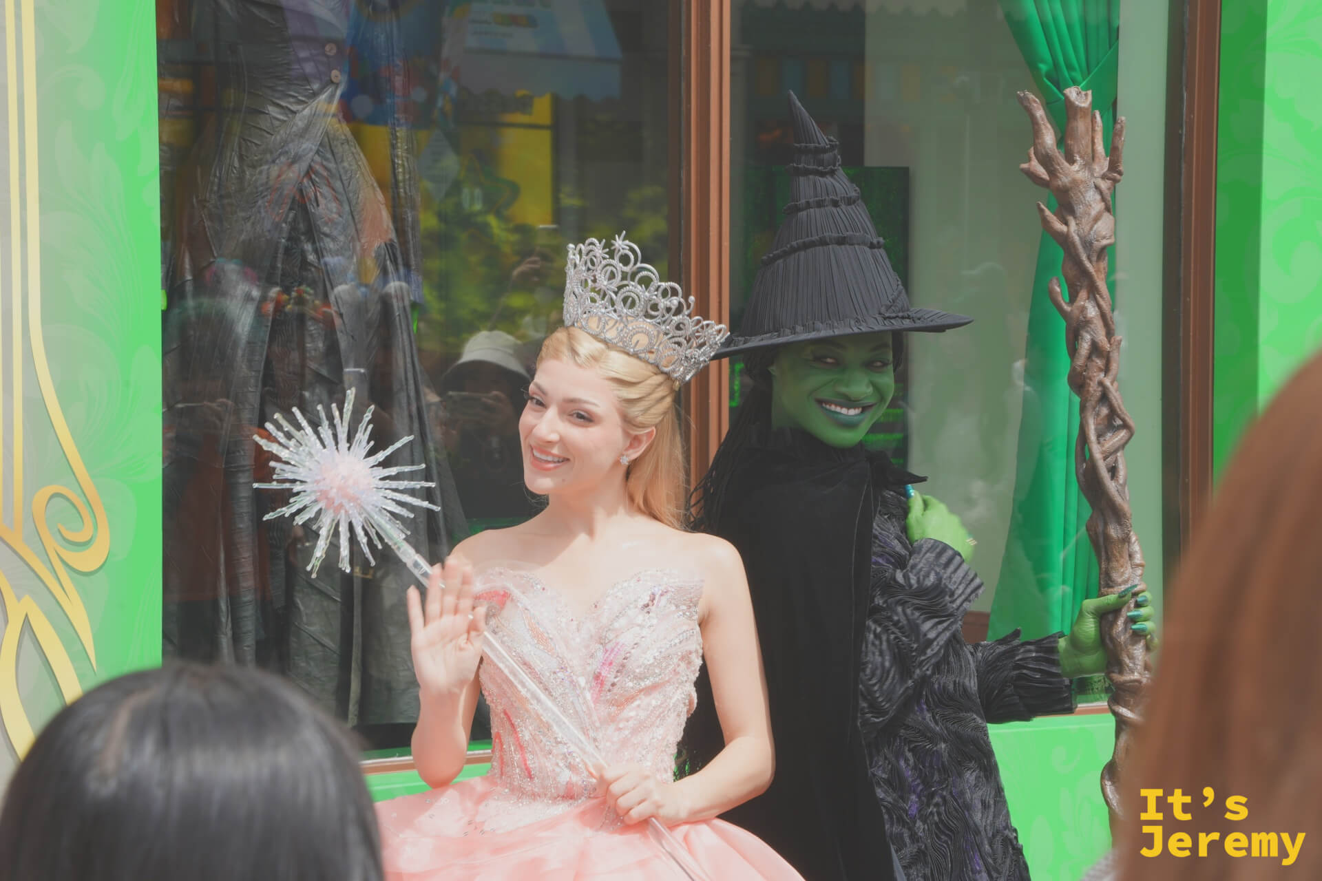 Glinda 與 Elphaba 並肩而立，一個穿著粉白蓬裙手持水晶魔法棒，另一個穿著黑色斗篷舉著老樹杖，形成強烈對比