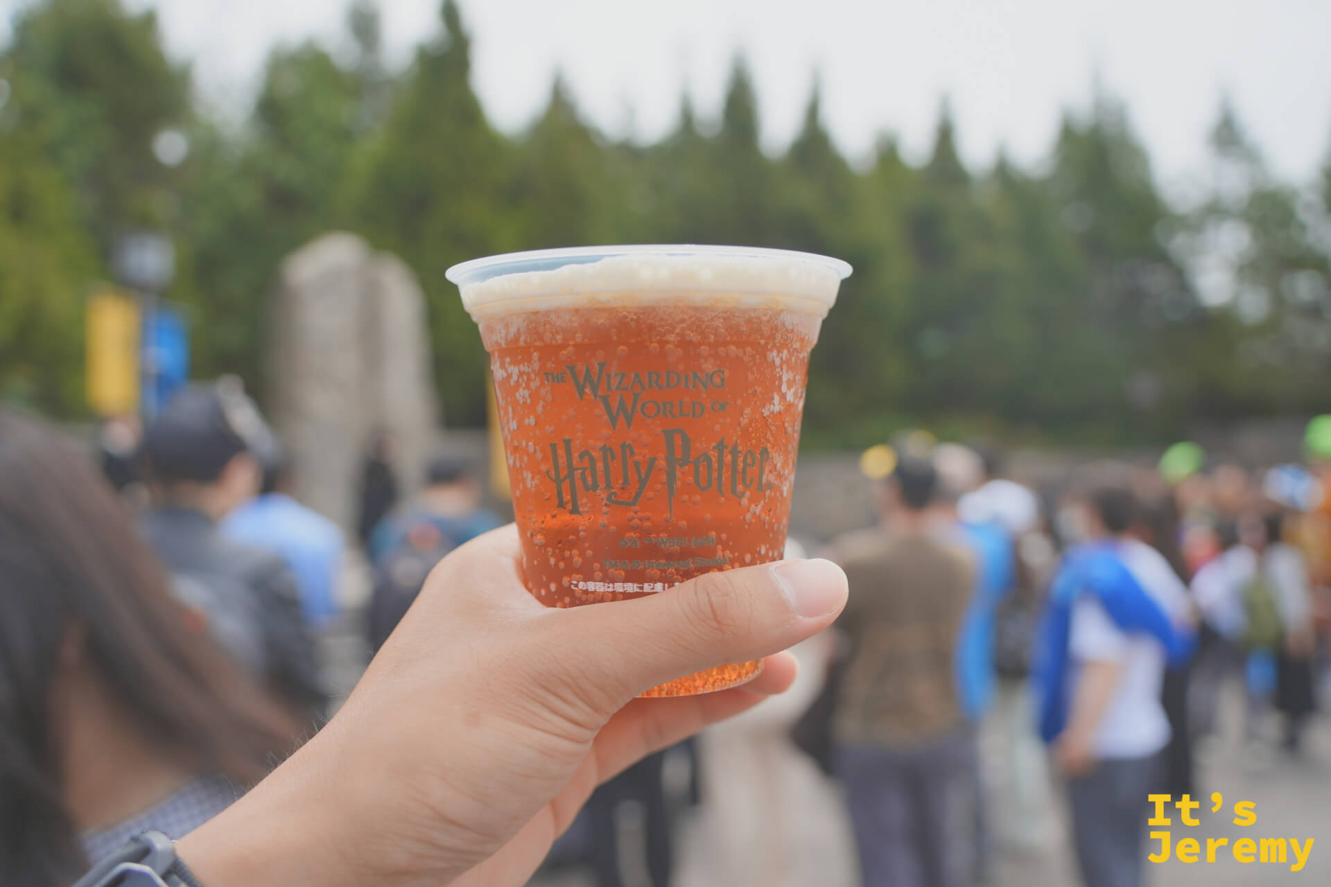 奶油啤酒，杯身印著「The Wizarding World of Harry Potter」字樣，橘紅色液體配上白色奶泡，在霍格莫德村的石牆前舉起來拍就很好看