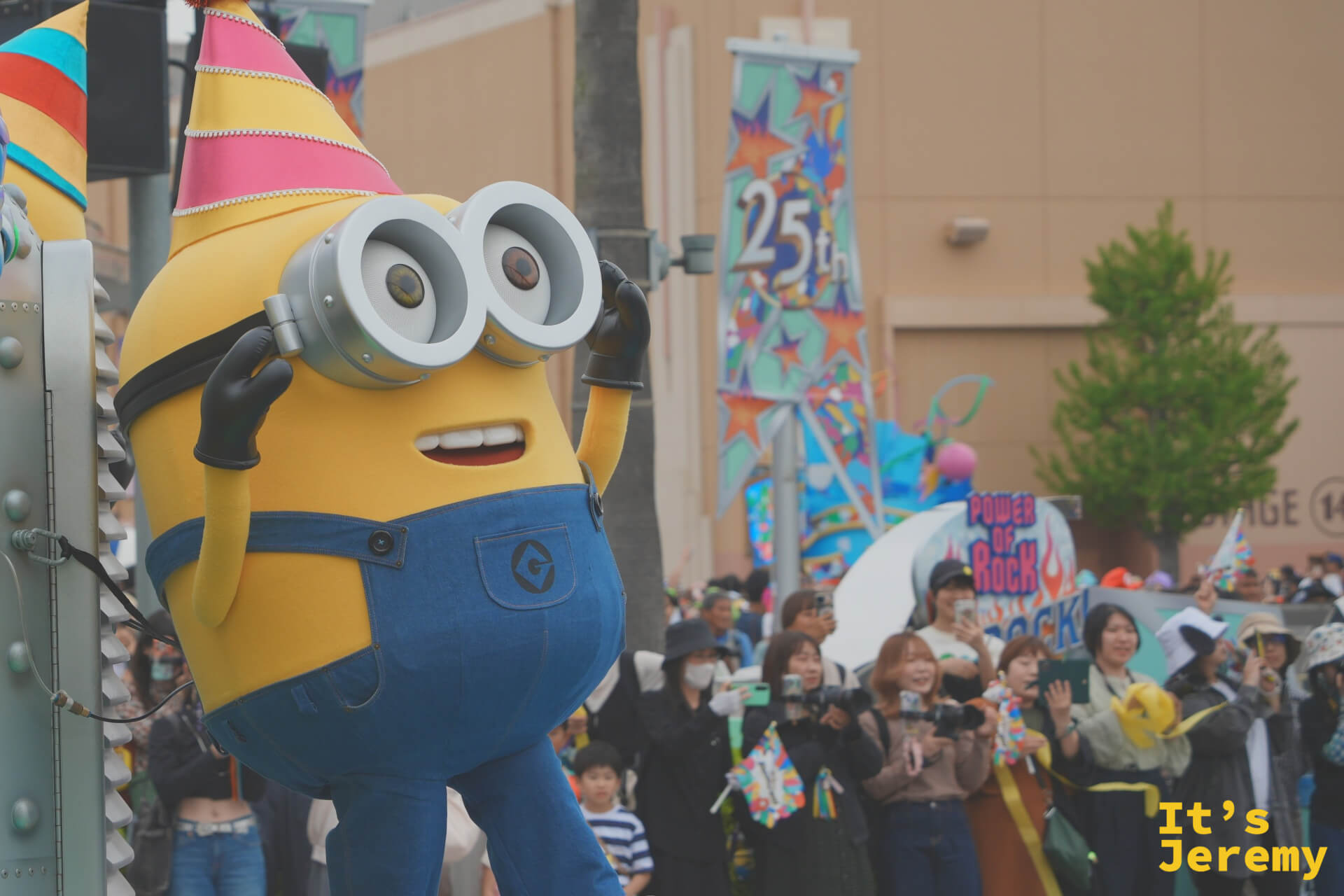 一隻 Minion 從花車旁的路燈基座探出身來，驚訝表情對著觀眾，25th 裝飾旗幟在背後飄揚