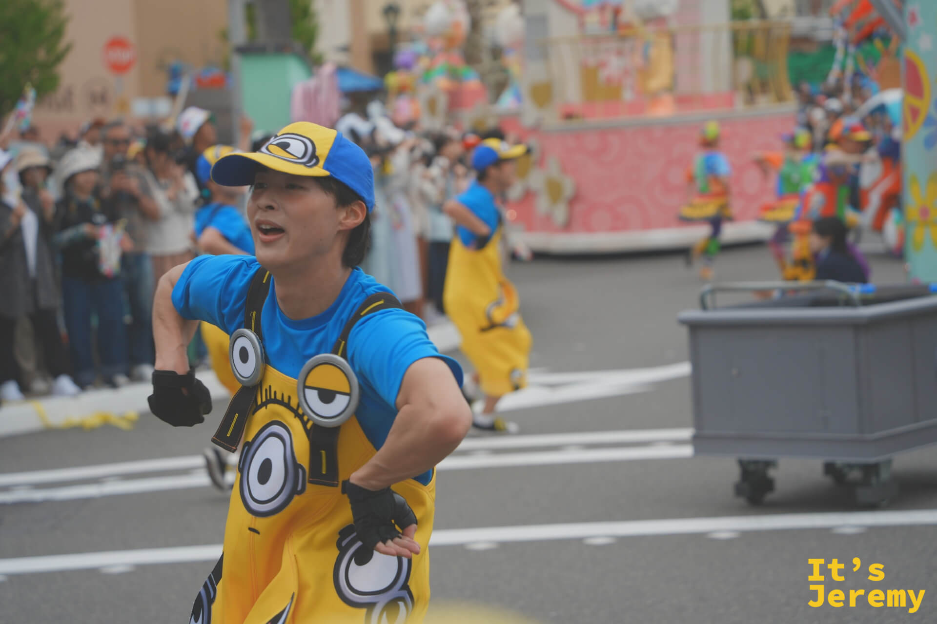 一位身穿 Minion 服裝的舞者在街道上奔跑，後方是色彩繽紛的遊行花車
