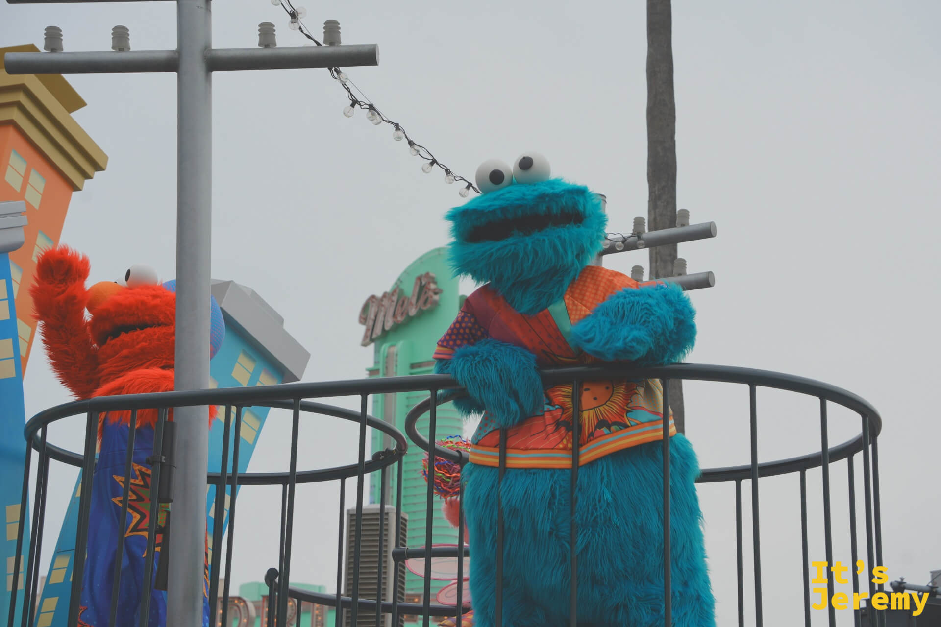 Cookie Monster 與 Elmo 並肩站在花車上，背景是芝麻街特色的彩色磚牆建築，遠處是 Mel
