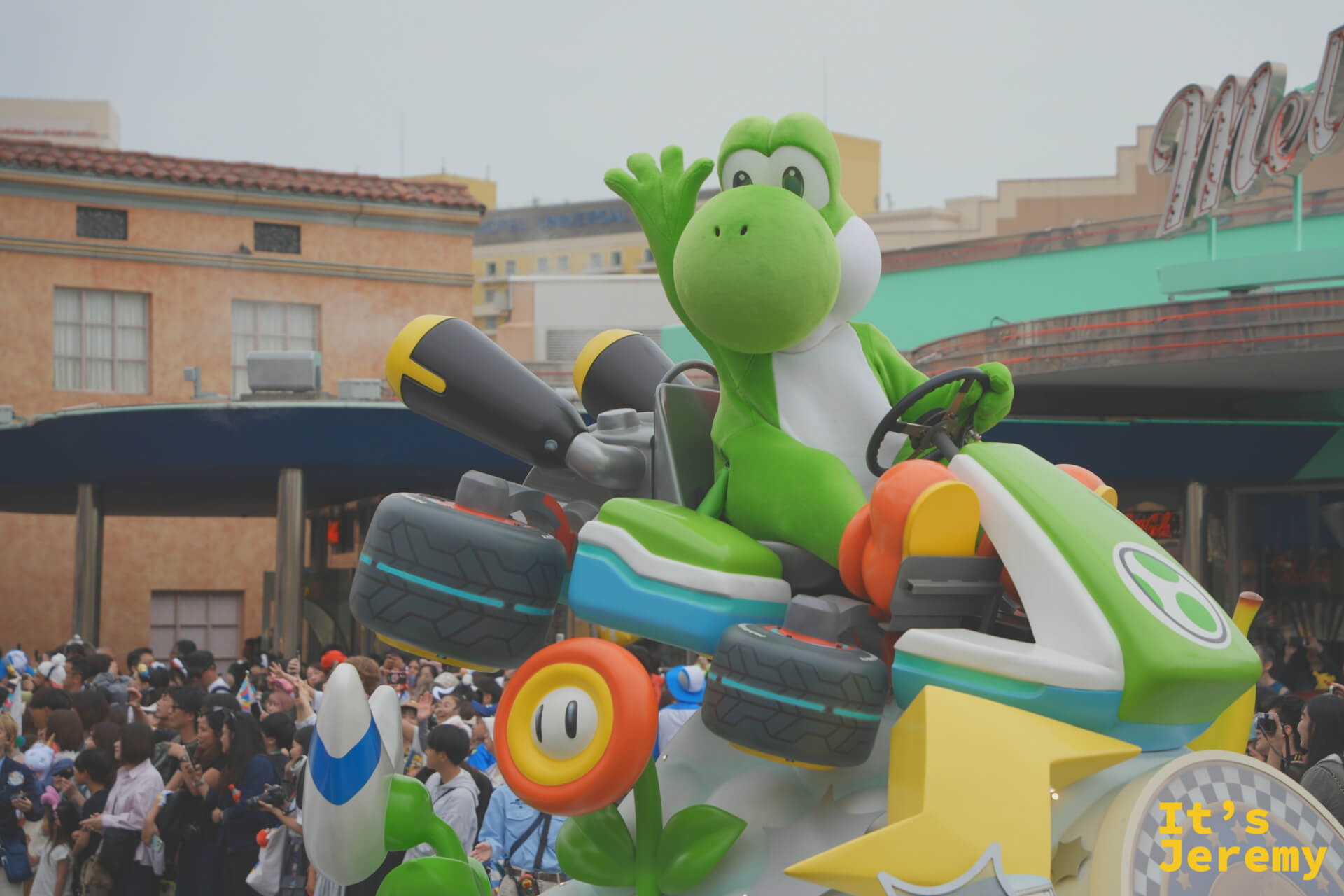Yoshi 特寫，綠色身軀坐在 Mario Kart 賽車裡，對著觀眾揮手，Mel