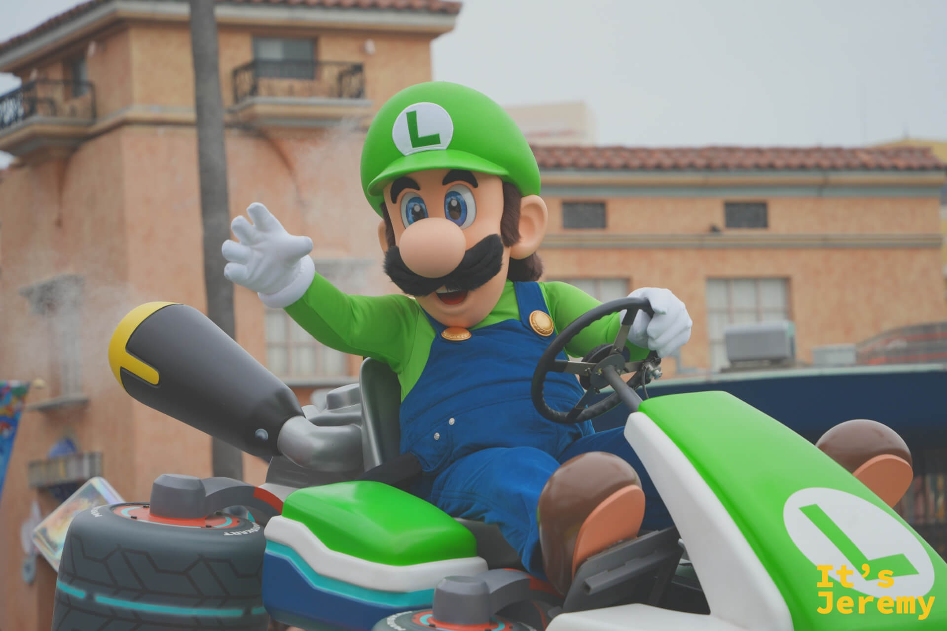Luigi 特寫，L 字賽車裡的 Luigi 對著攝影機揮手，後方的 Mel