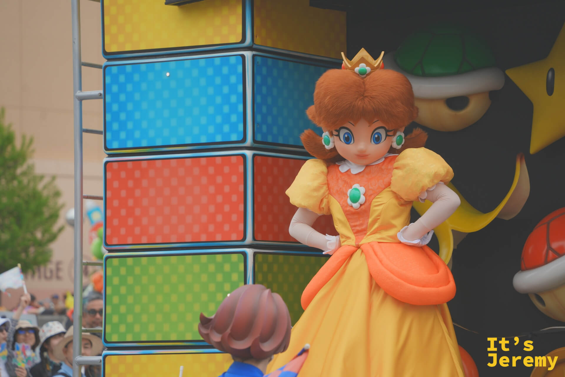 Princess Daisy 單獨特寫，橘黃蓬裙配上 D 字項鍊，站在馬賽克磚塊花車邊沿，角落還隱約可見一顆 Poké Ball
