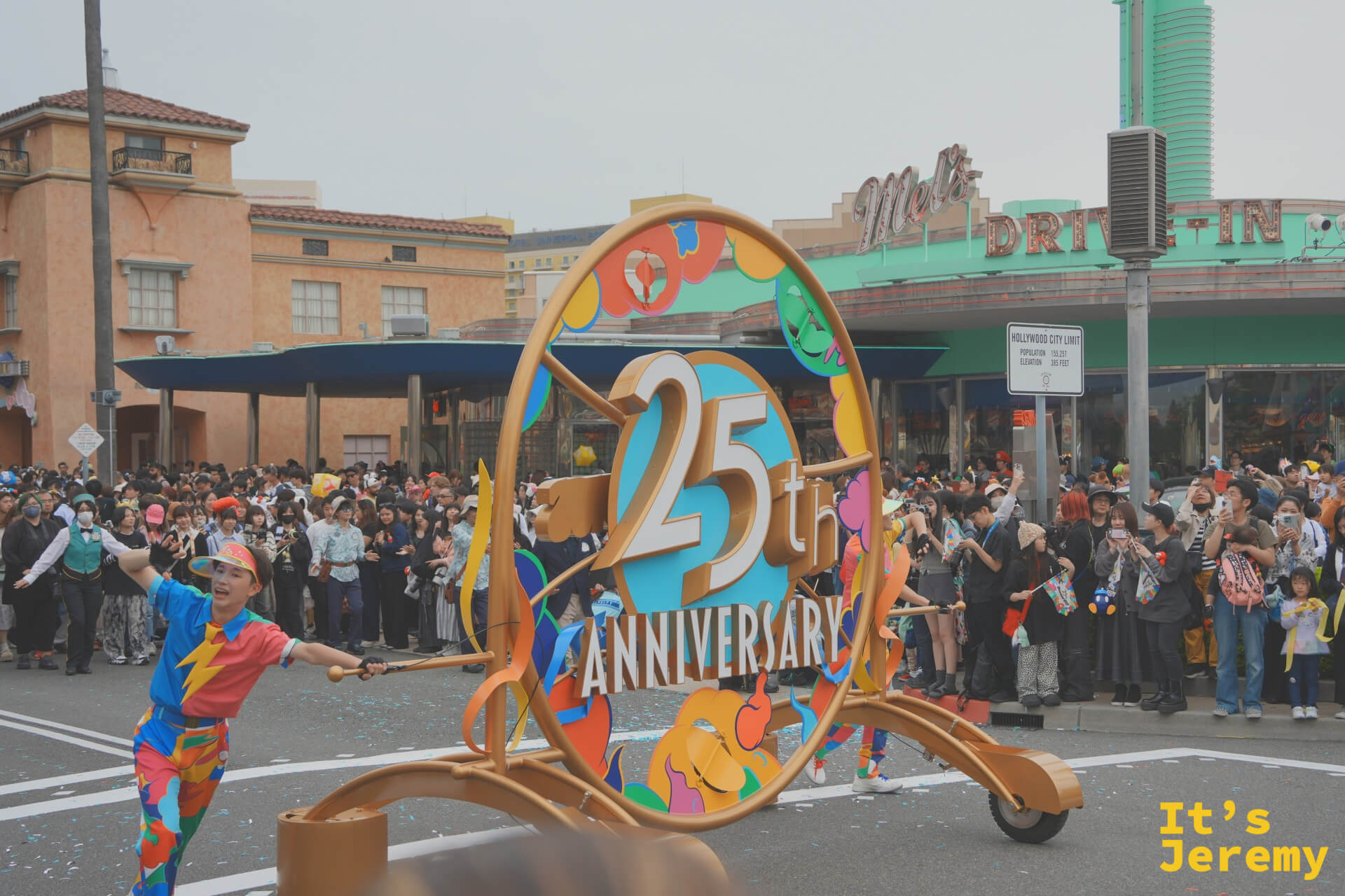 25th Anniversary 金色圓形標誌特寫，演員在兩側舉著標誌在街道上走動
