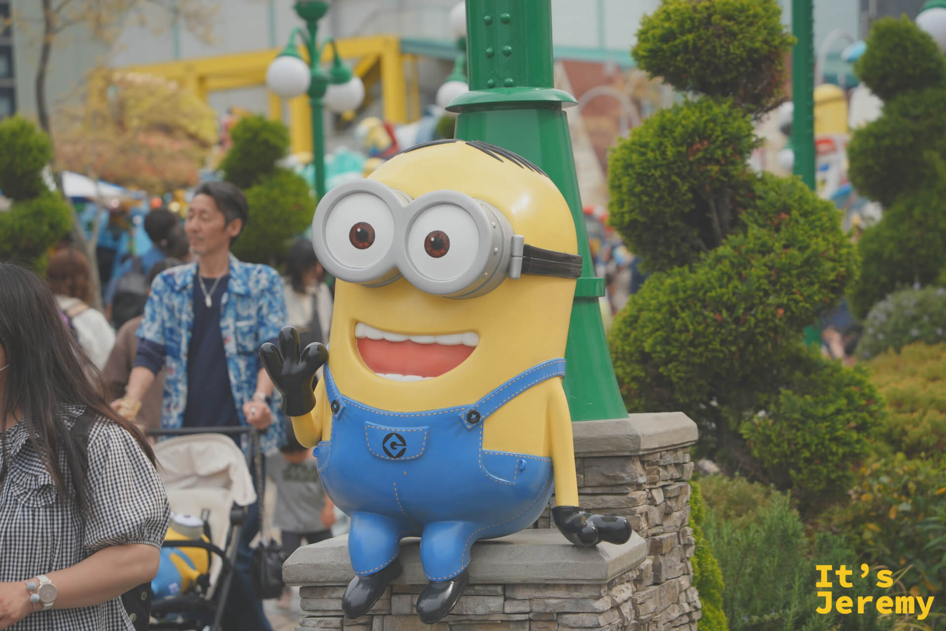 Minion 雕像坐在庭院的低矮石墩上，背後是黃綠相間的 Minion Park 景觀