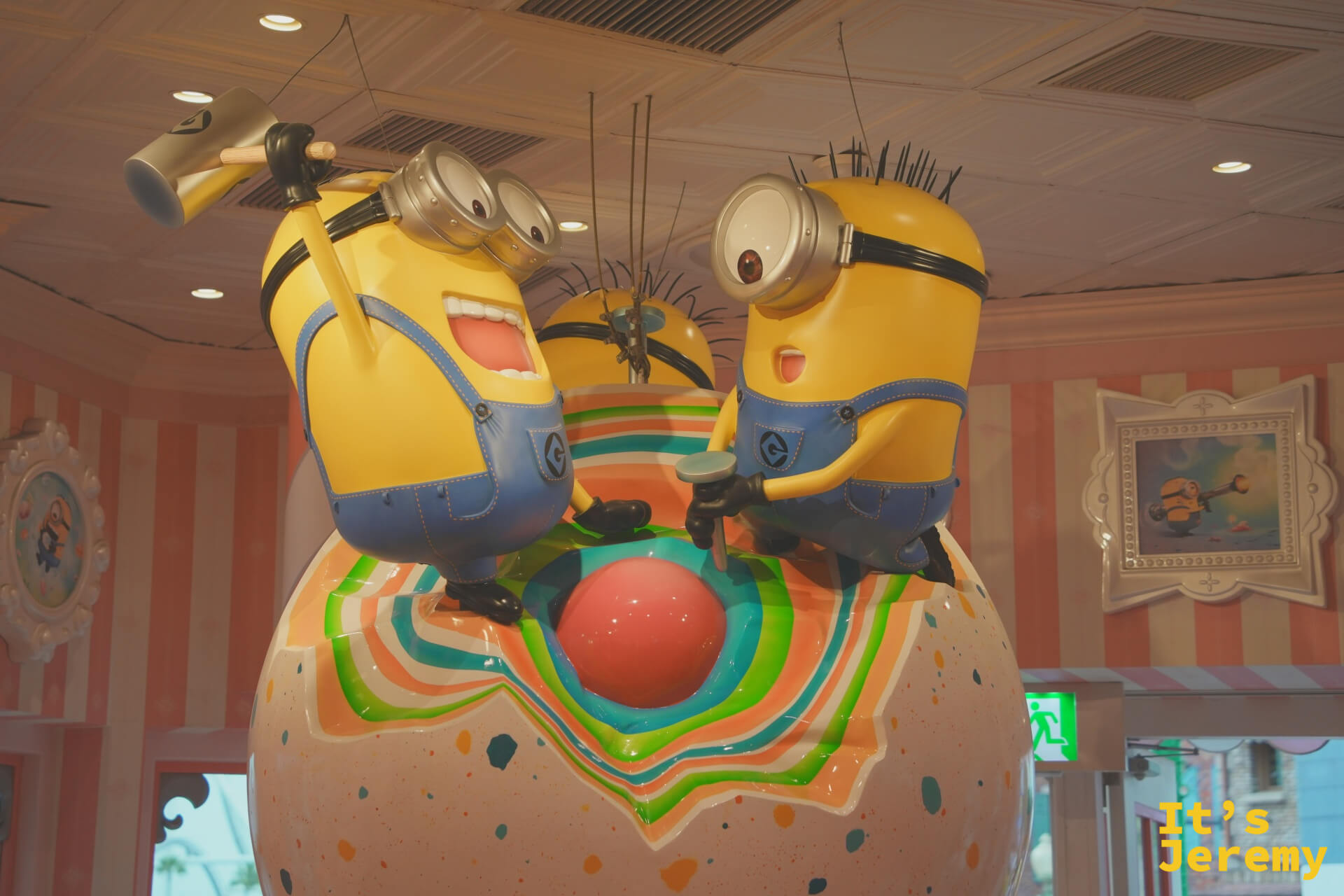 三隻 Minion 雕像攀在甜甜圈球體上，表情各異，從天花板俯瞰整個粉色系糕點風格店內