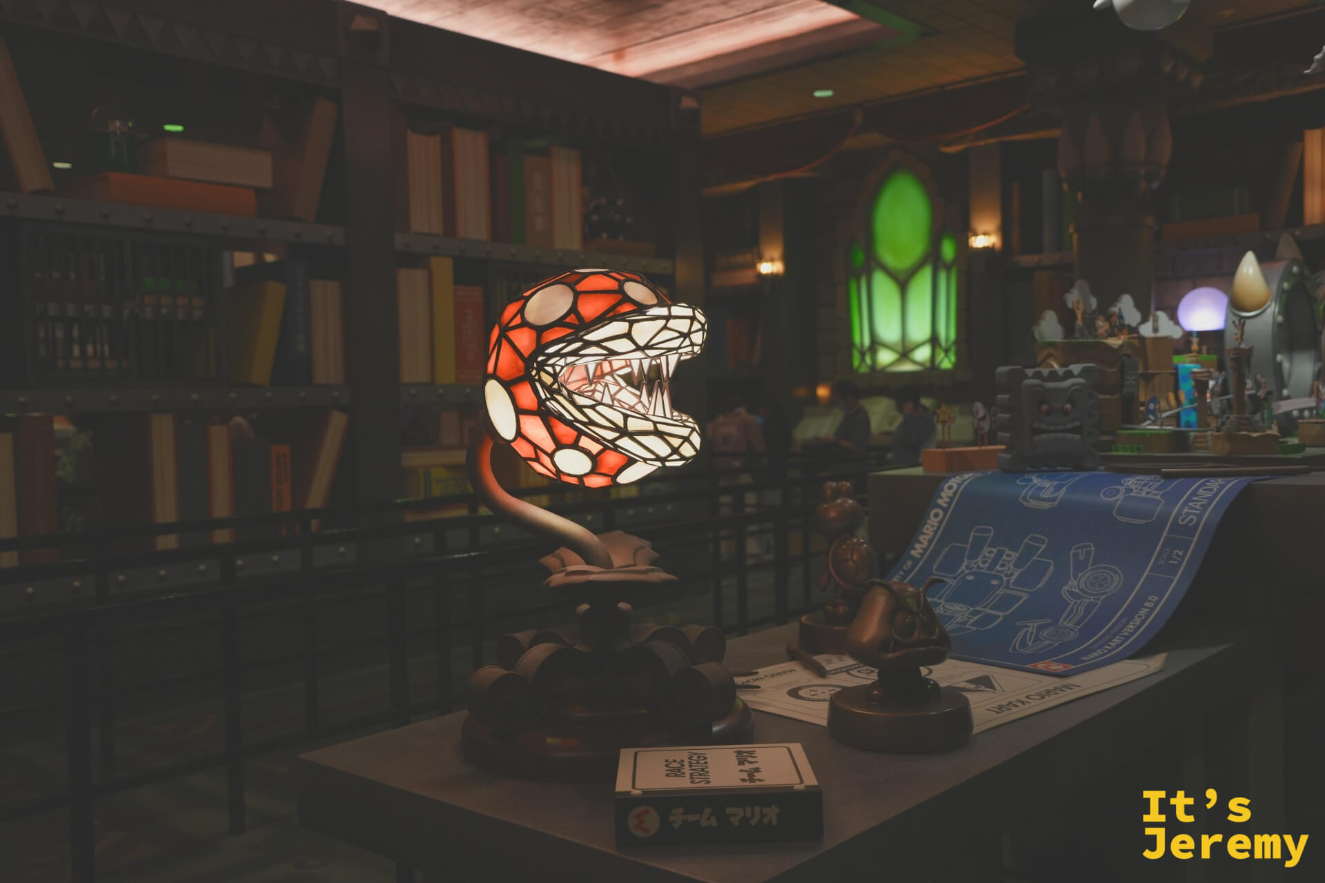 SNW 商店內的 Piranha Plant（吃人花）彩繪玻璃台燈，旁邊散落著 Mario Motocross 設計圖，整個區域的道具設計充滿細節