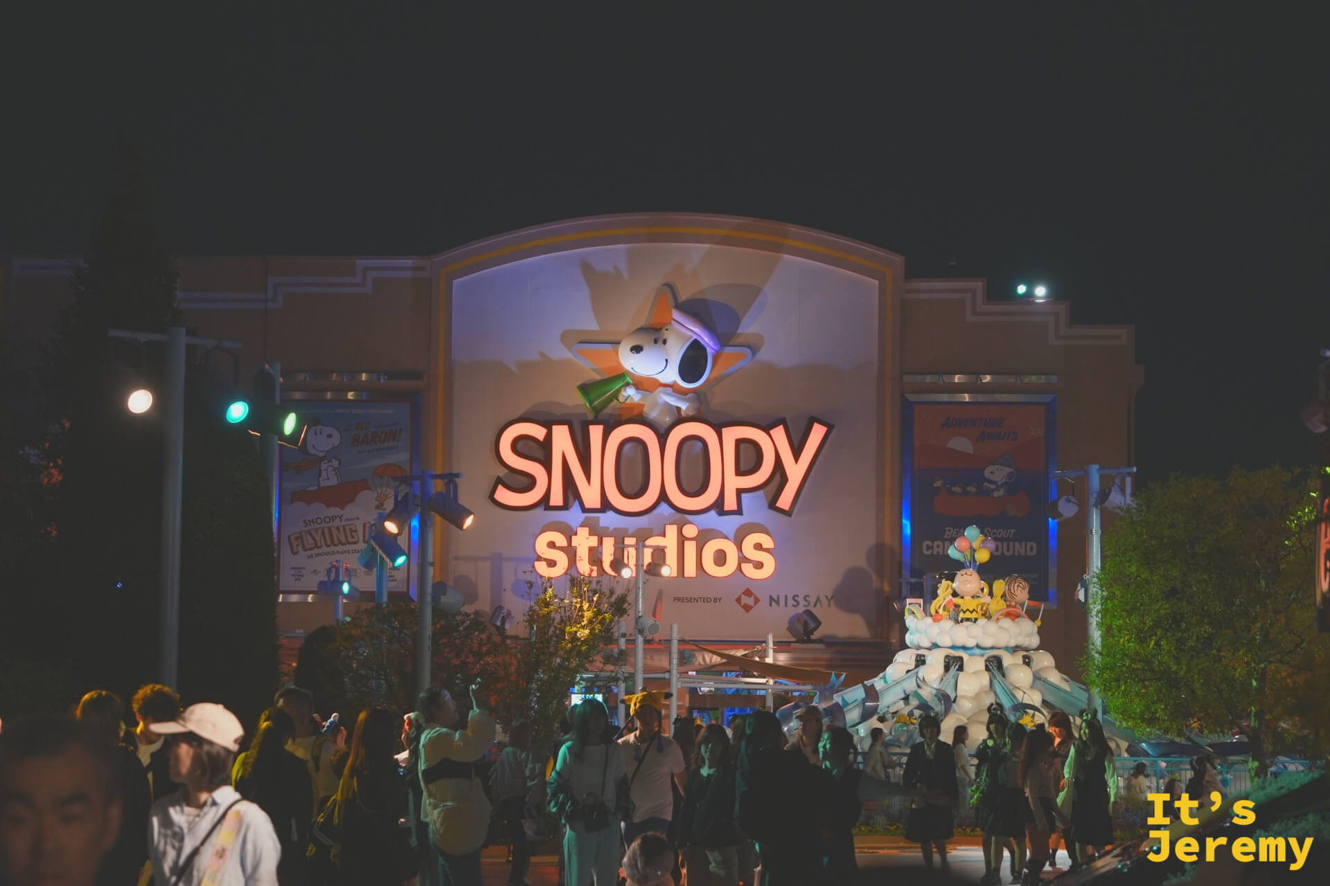 Snoopy Studios 夜景，霓虹燈管組成的 Snoopy 圖案在建築正面閃爍，旁邊有 25 週年白色蛋糕裝置，入口廣場人潮絡繹不絕