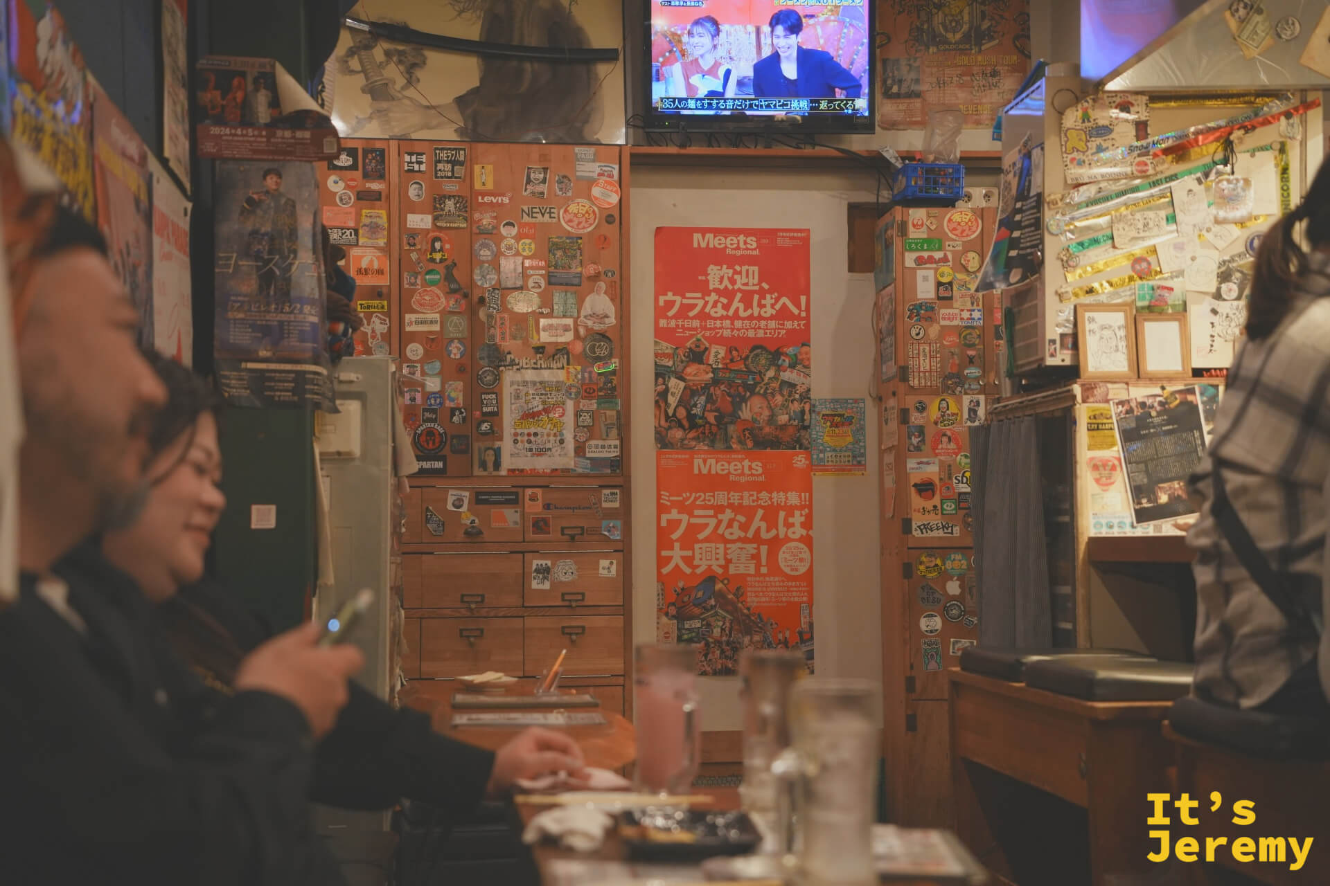 客人在吧台前聊天大笑，背後貼著「歡迎，ウラなんばへ！」Meets 雜誌海報，電視在角落播放