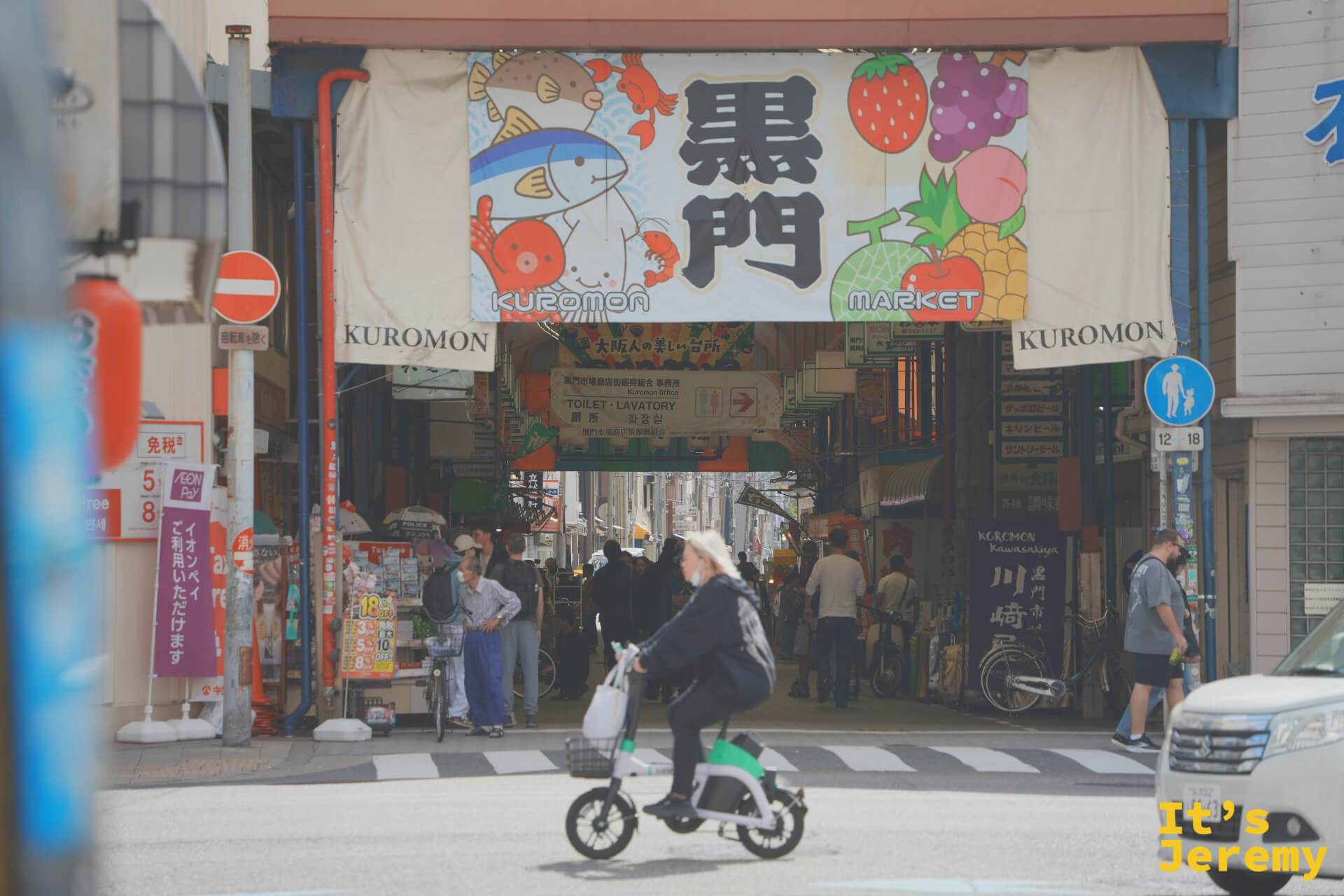 市場入口近拍，招牌下騎腳踏車的行人與周邊店家