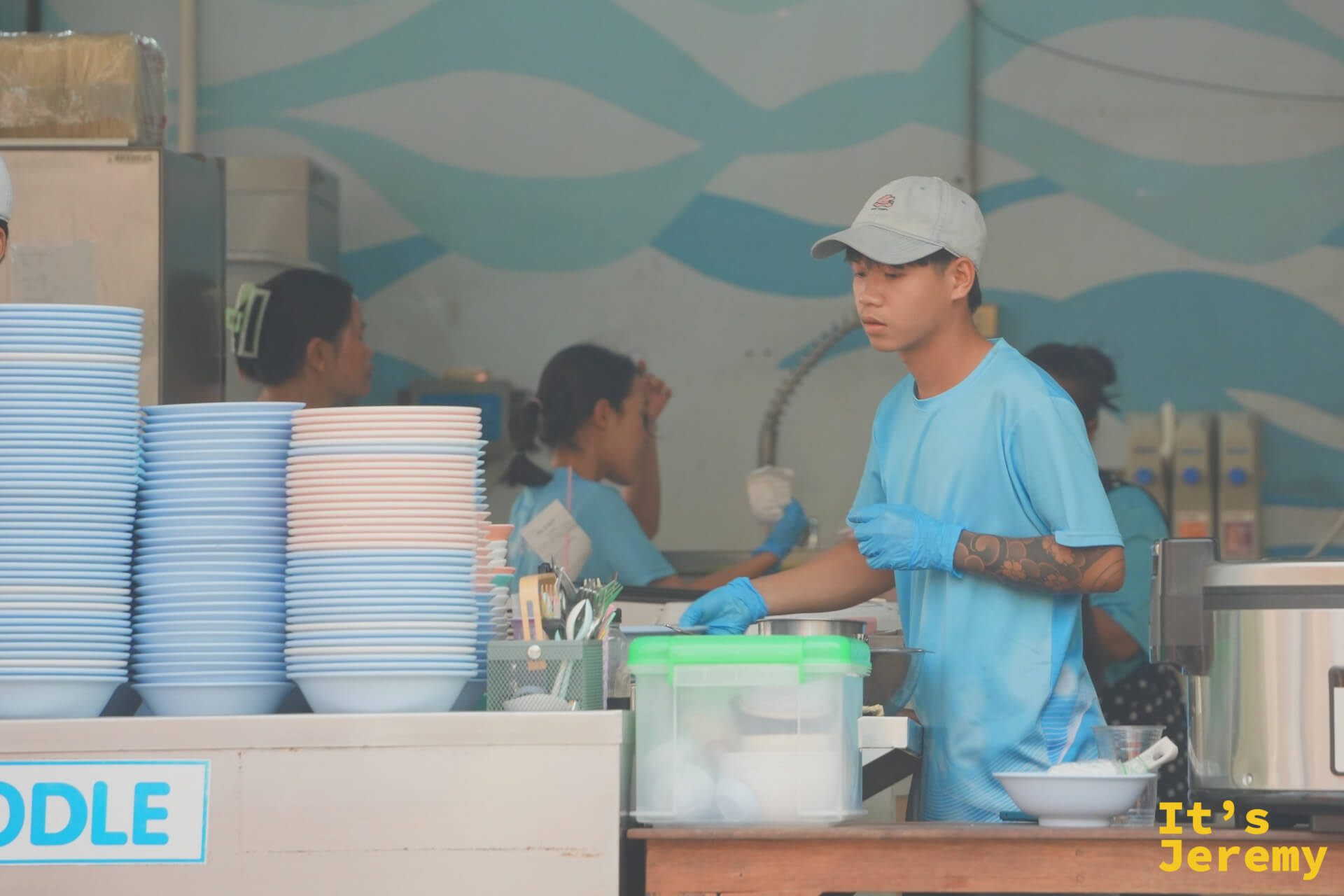 廚房工作人員特寫，戴著藍色手套備餐，背後是海浪壁畫