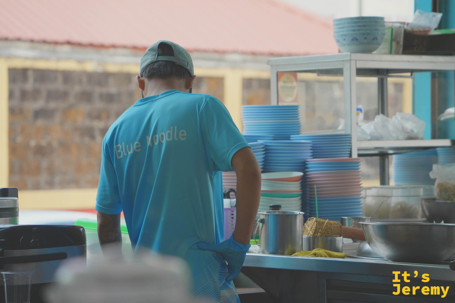 穿 Blue Noodle 制服的男店員在廚房備餐，背後是整齊堆疊的藍色碗盤